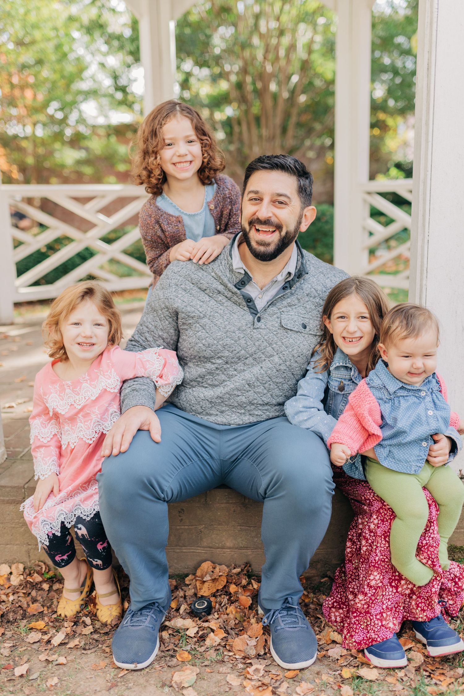 A smiling man sits outdoors with four young girls surrounding him, posing for a cheerful family portrait in autumn.