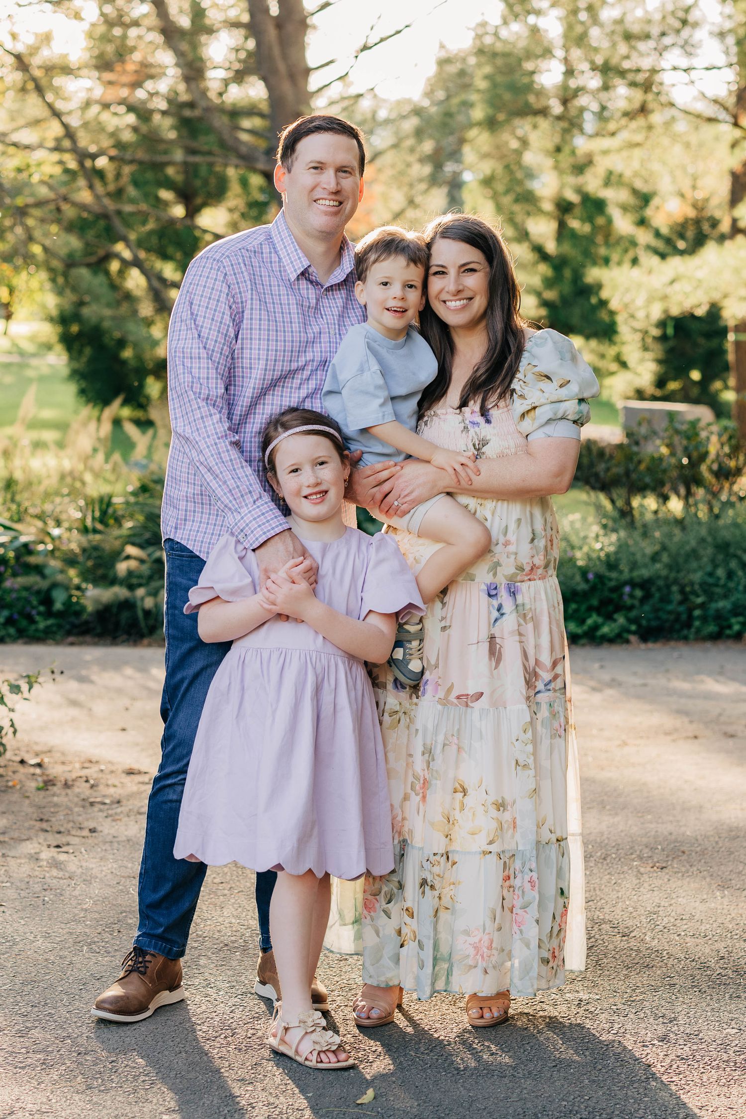A happy family of four poses outdoors, parents with two young children in pastel outfits, surrounded by lush greenery.