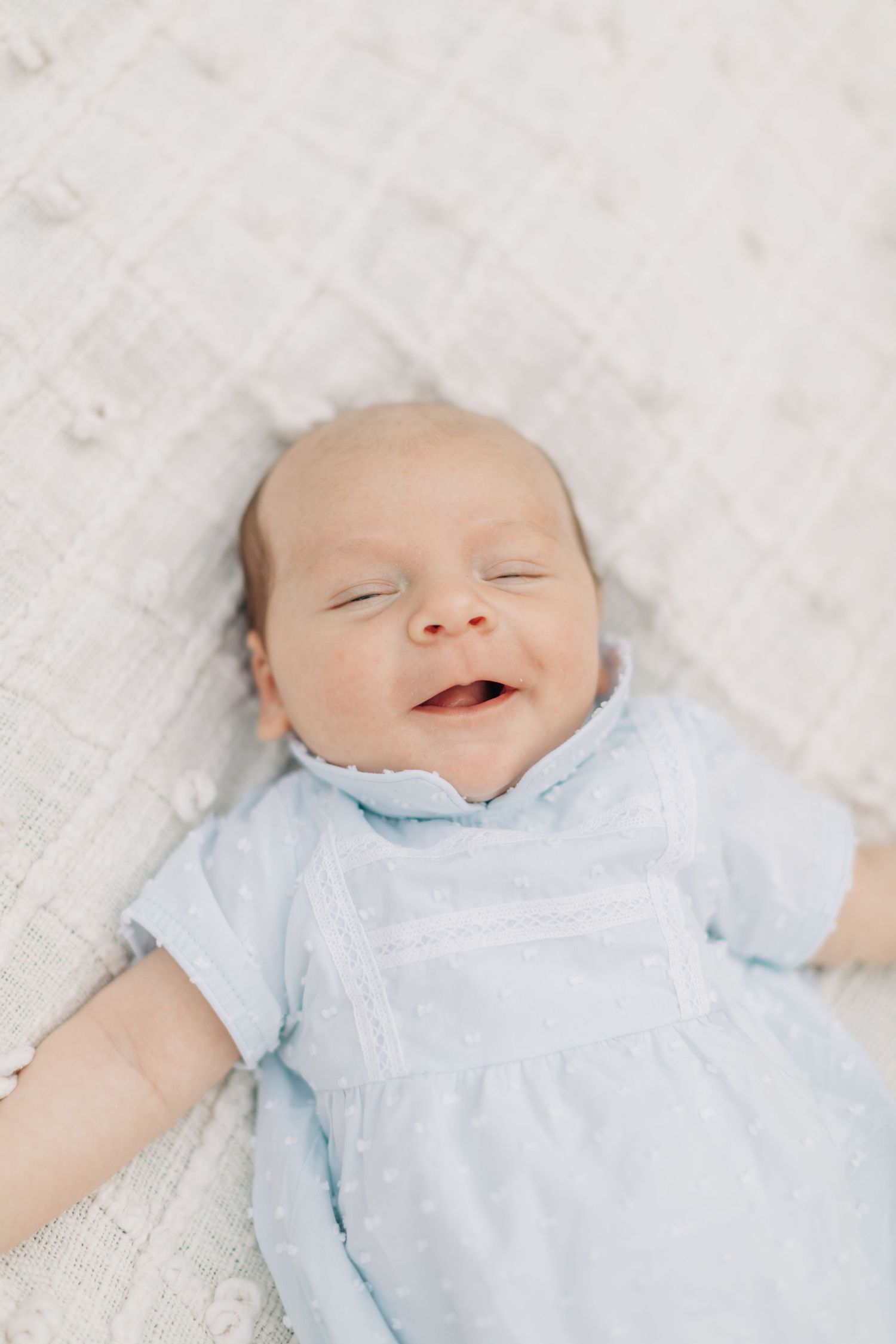 Smiling baby in light blue outfit on white blanket.