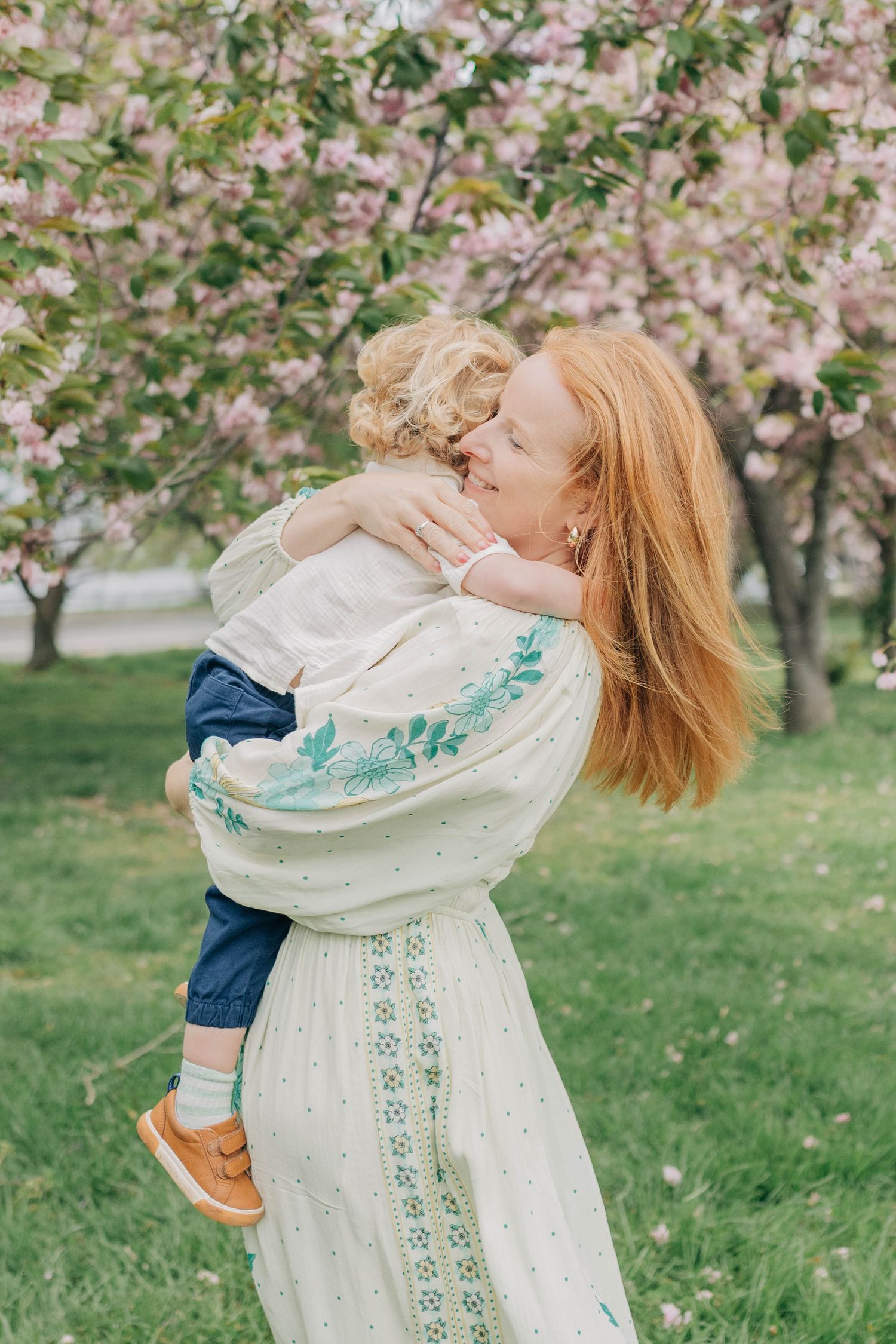 A tender embrace under blooming cherry blossoms, with a white embroidered dress and spring greenery in the background.