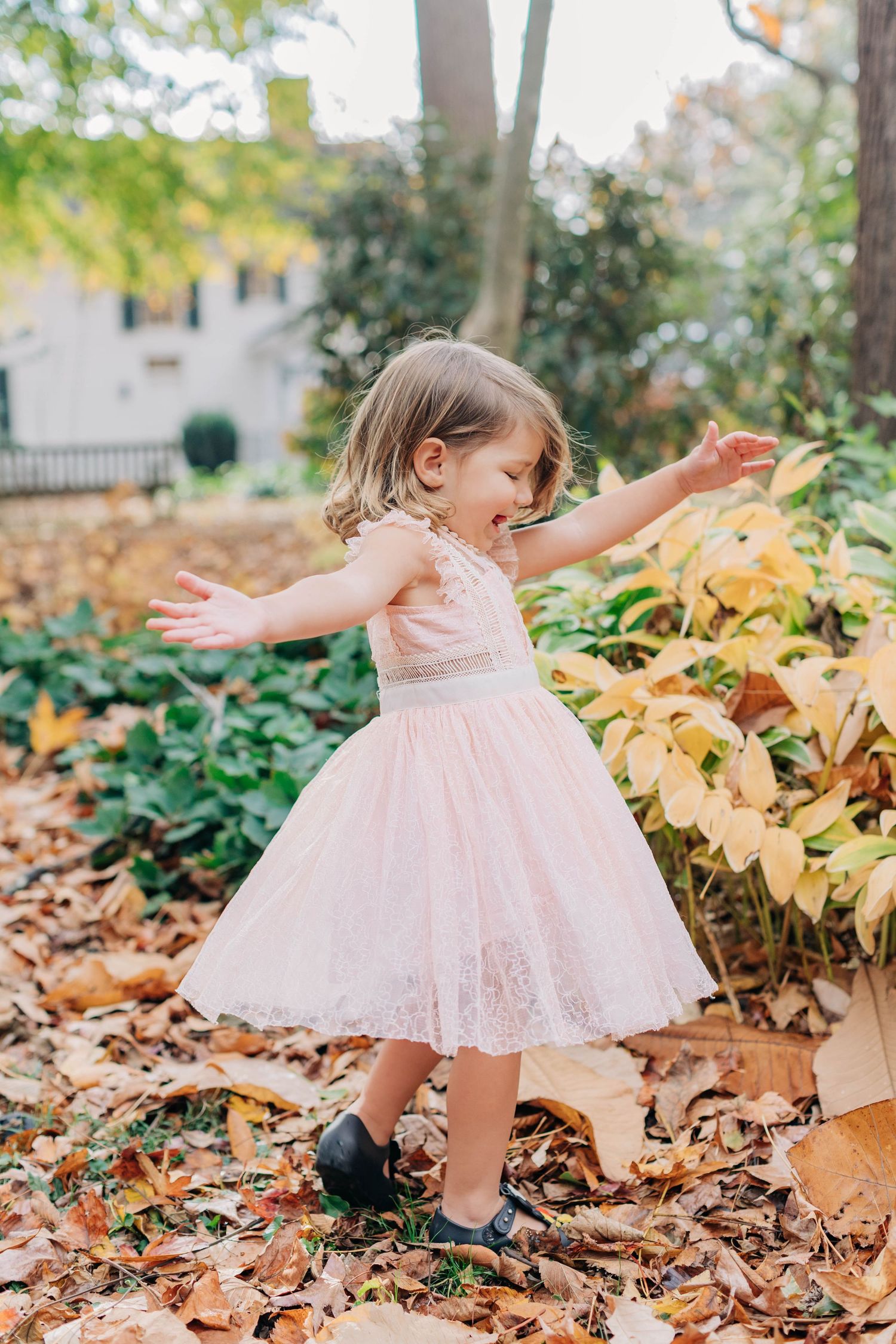 Child in pink dress twirling among fallen autumn leaves during outdoor photoshoot.