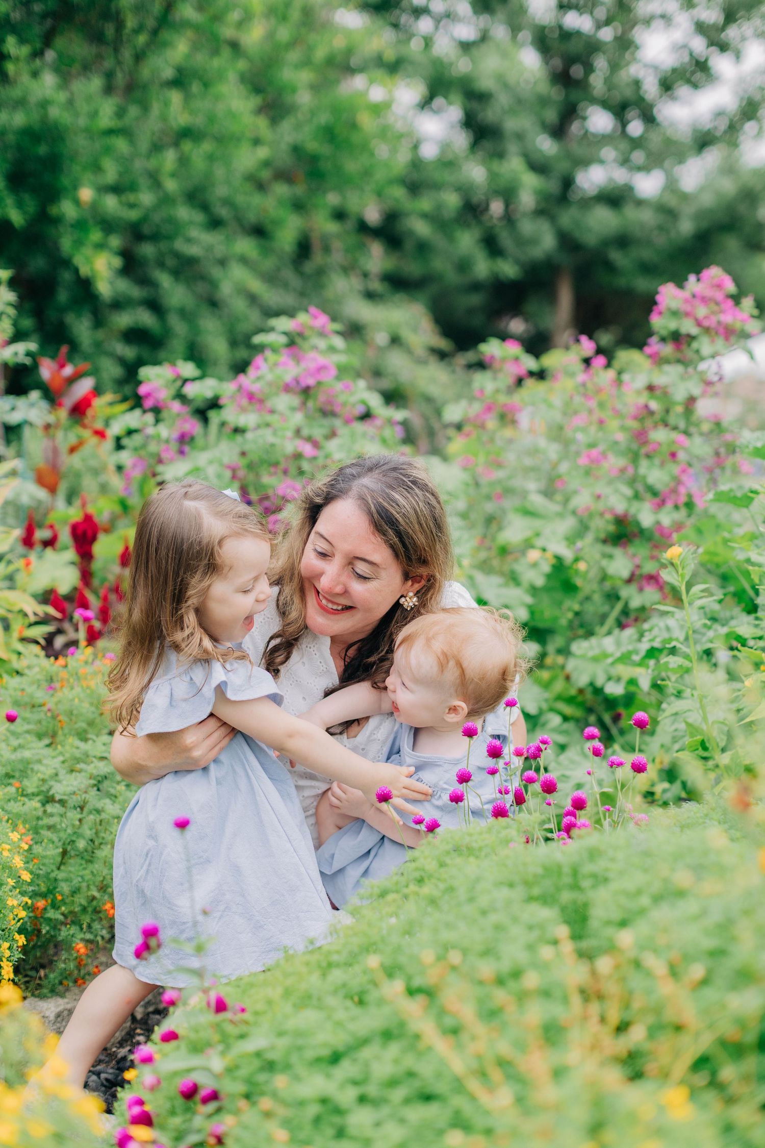 A mother smiles while holding two young children in a lush garden filled with colorful blooming flowers.