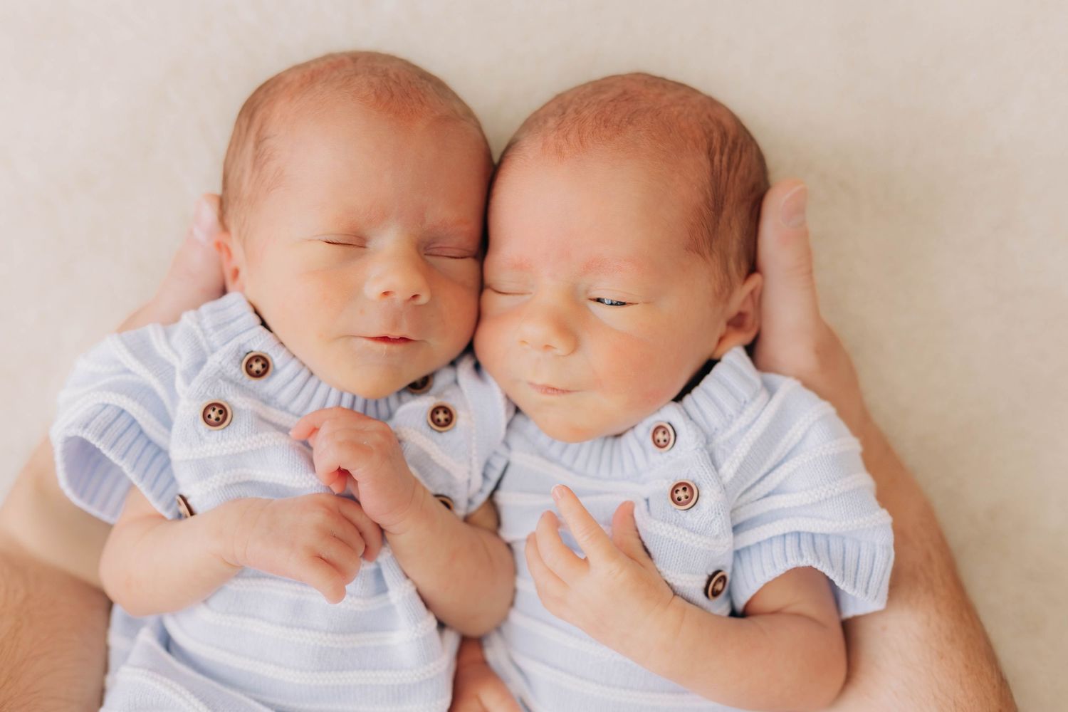 Two sleeping newborn twins in light blue striped rompers lying side by side on a soft cream blanket.