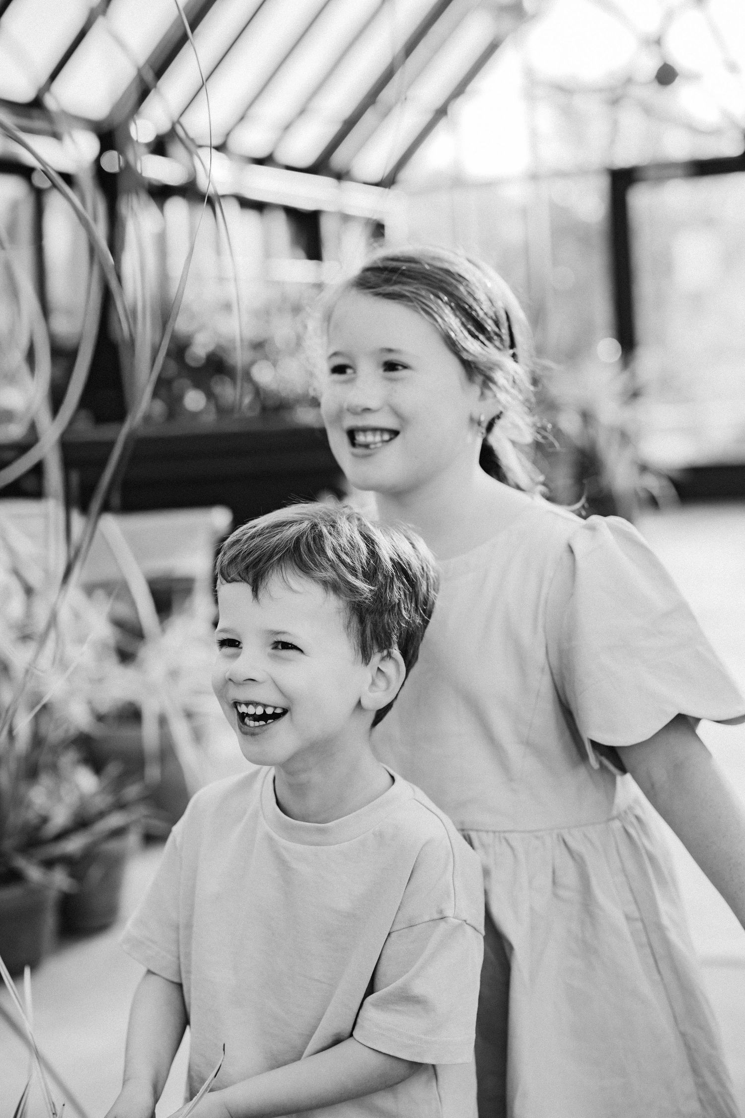 A black and white photo of a smiling boy and girl standing together in a greenhouse.