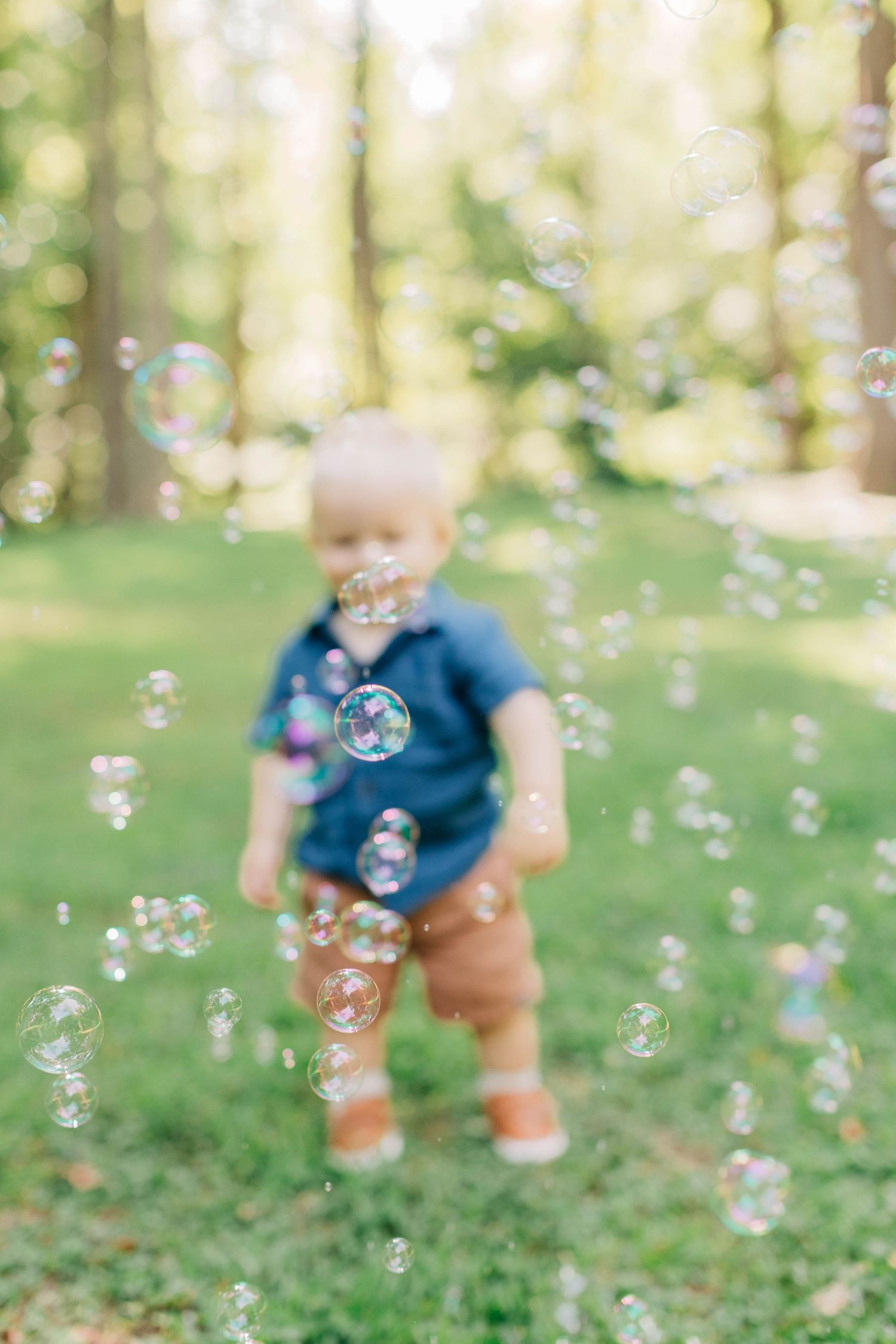 A curious toddler in blue shirt and brown pants plays with floating soap bubbles in a sunny park.