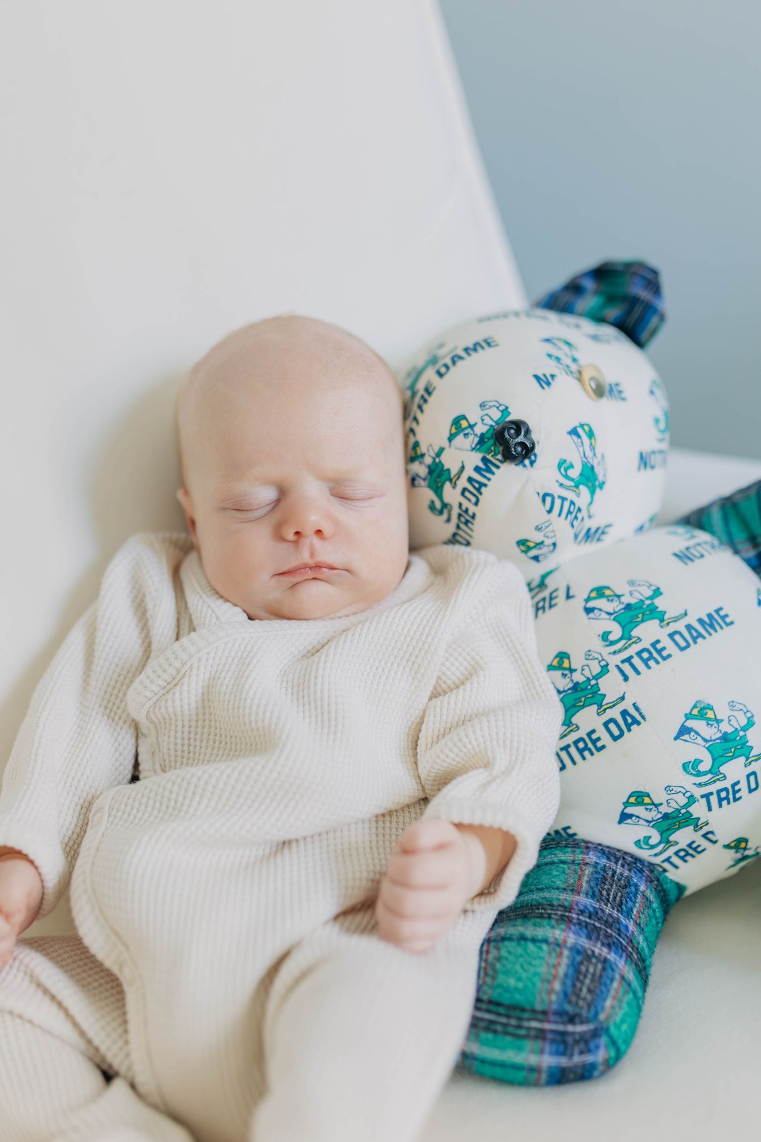 Peaceful newborn baby sleeping in white outfit beside Notre Dame themed teddy bear.