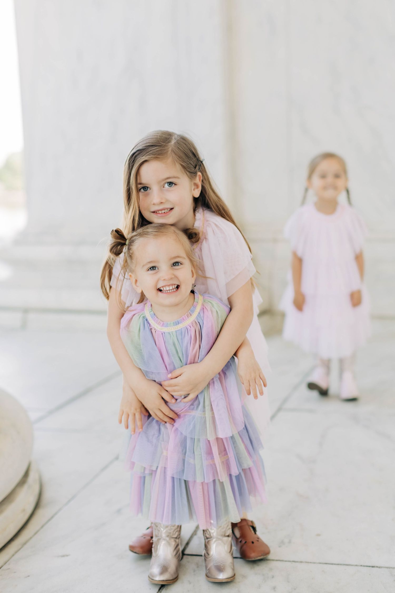Two little girls in pastel dresses embrace warmly while a third girl stands softly blurred in the background.