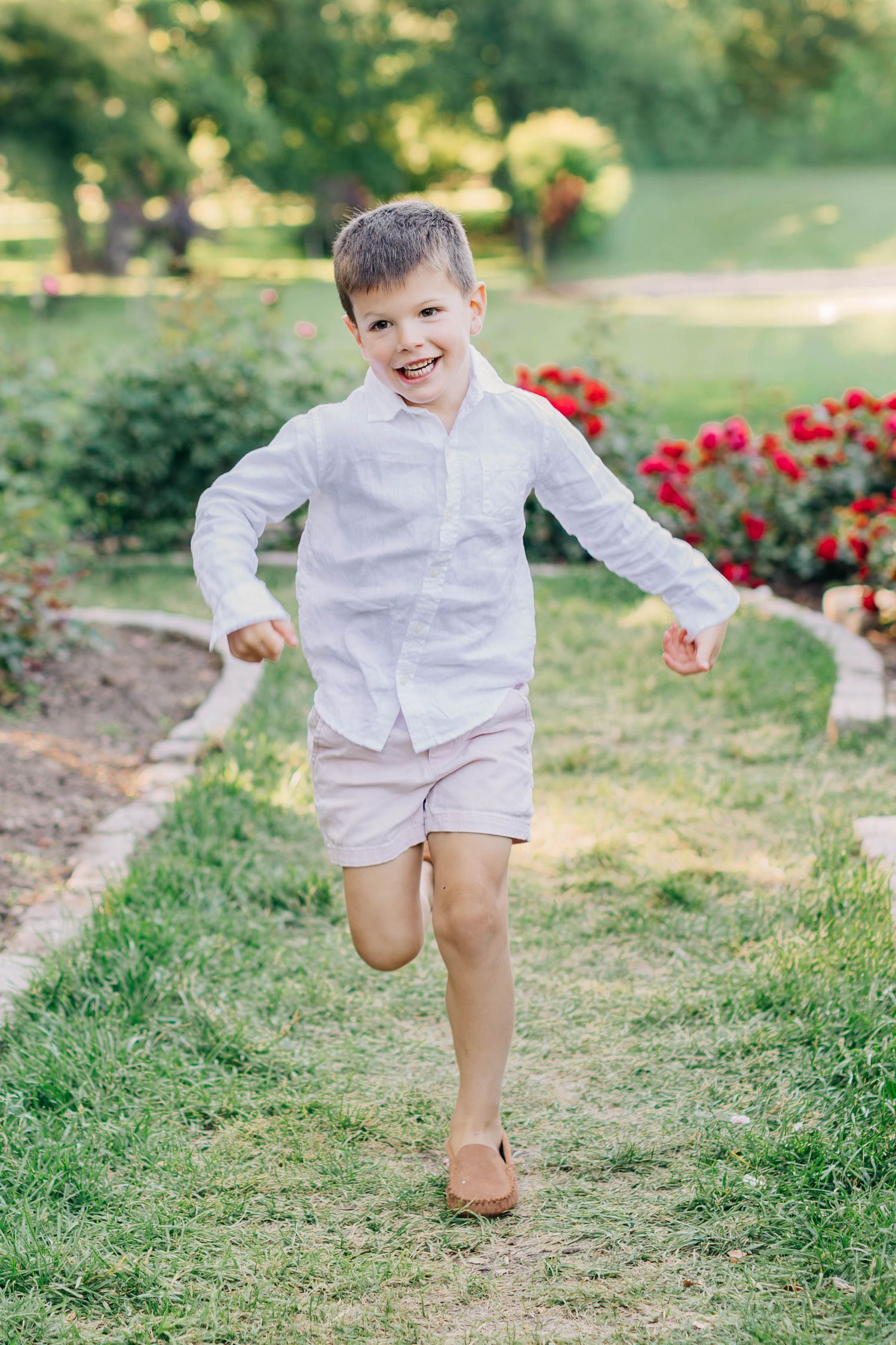 Happy young boy in white shirt and khaki shorts running and smiling in a lush green garden.