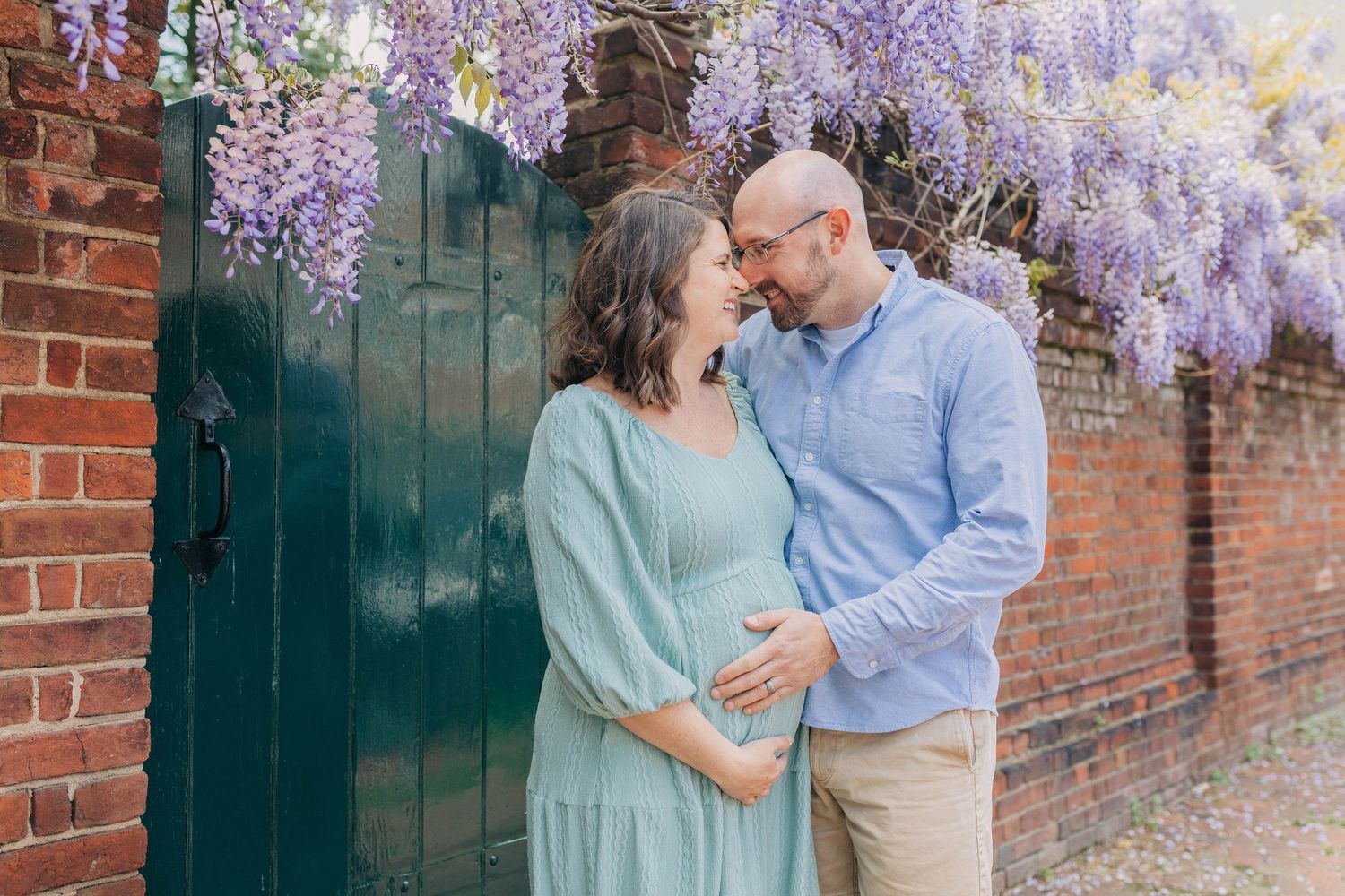 Expectant couple smiling at each other under purple wisteria by a teal gate and brick wall.