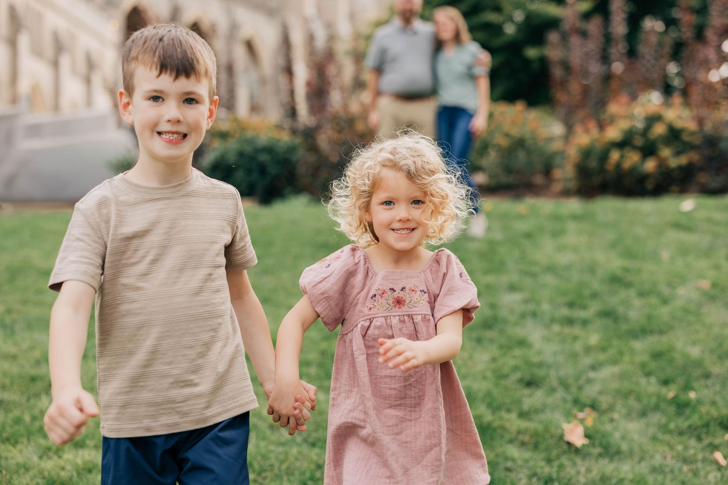 Two smiling young children holding hands and walking across a sunny green lawn, with two adults standing in the background.