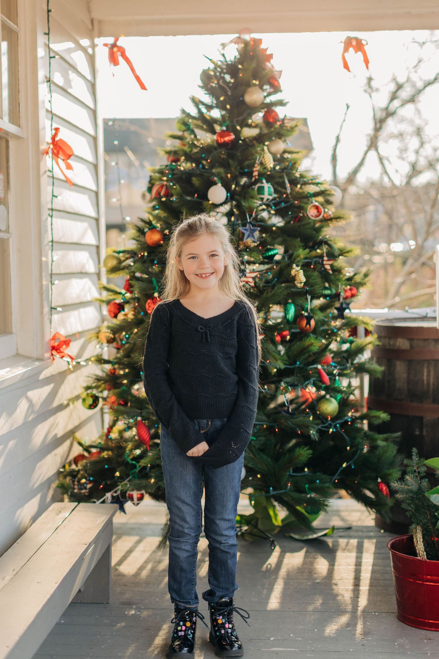 Young blonde girl smiling in front of decorated Christmas tree on porch, wearing black sweater and jeans.
