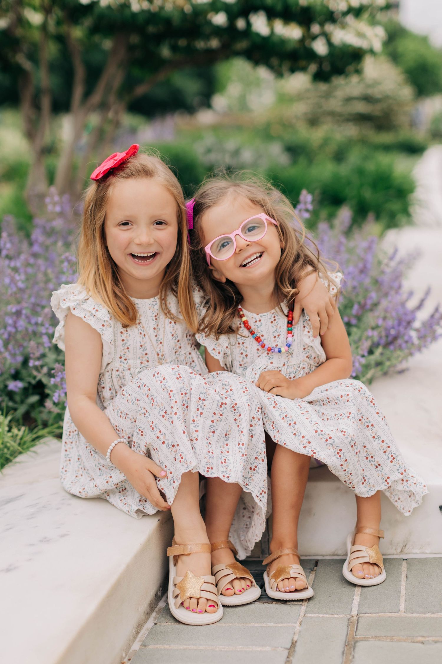 Two young girls in matching floral dresses smile together outdoors among purple lavender flowers.