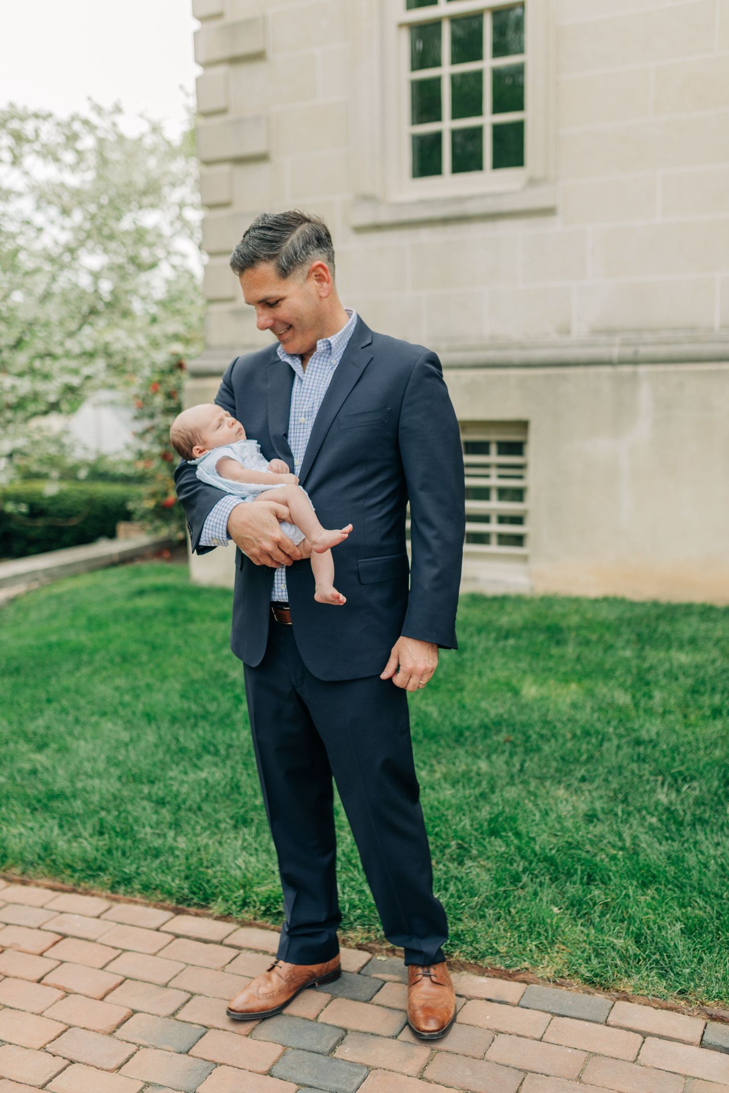 A tender moment captured outdoors as a newborn baby is held against a navy suit on a brick pathway.