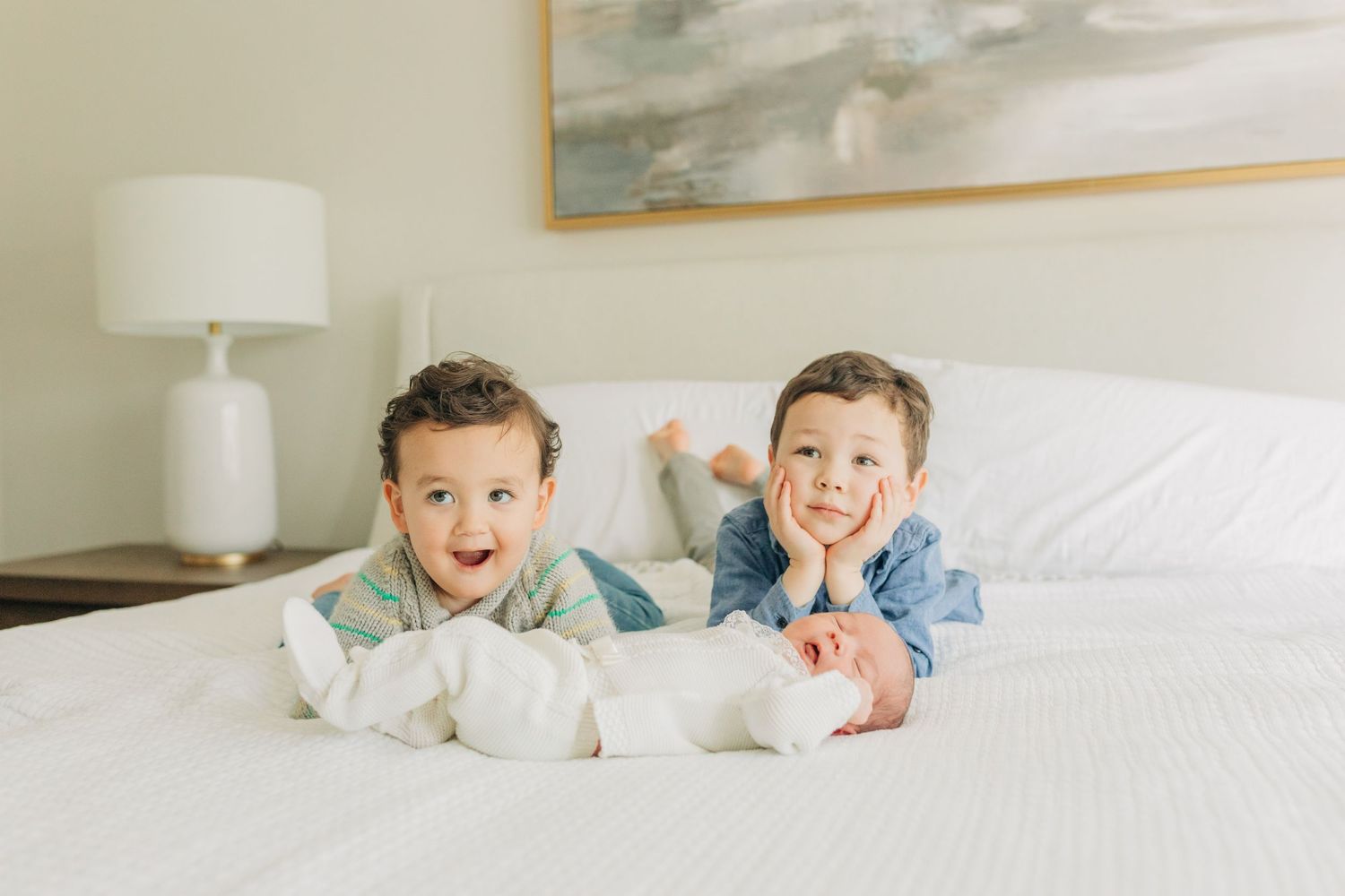 Children laying on white bedding showing happy expressions with artwork visible in background.