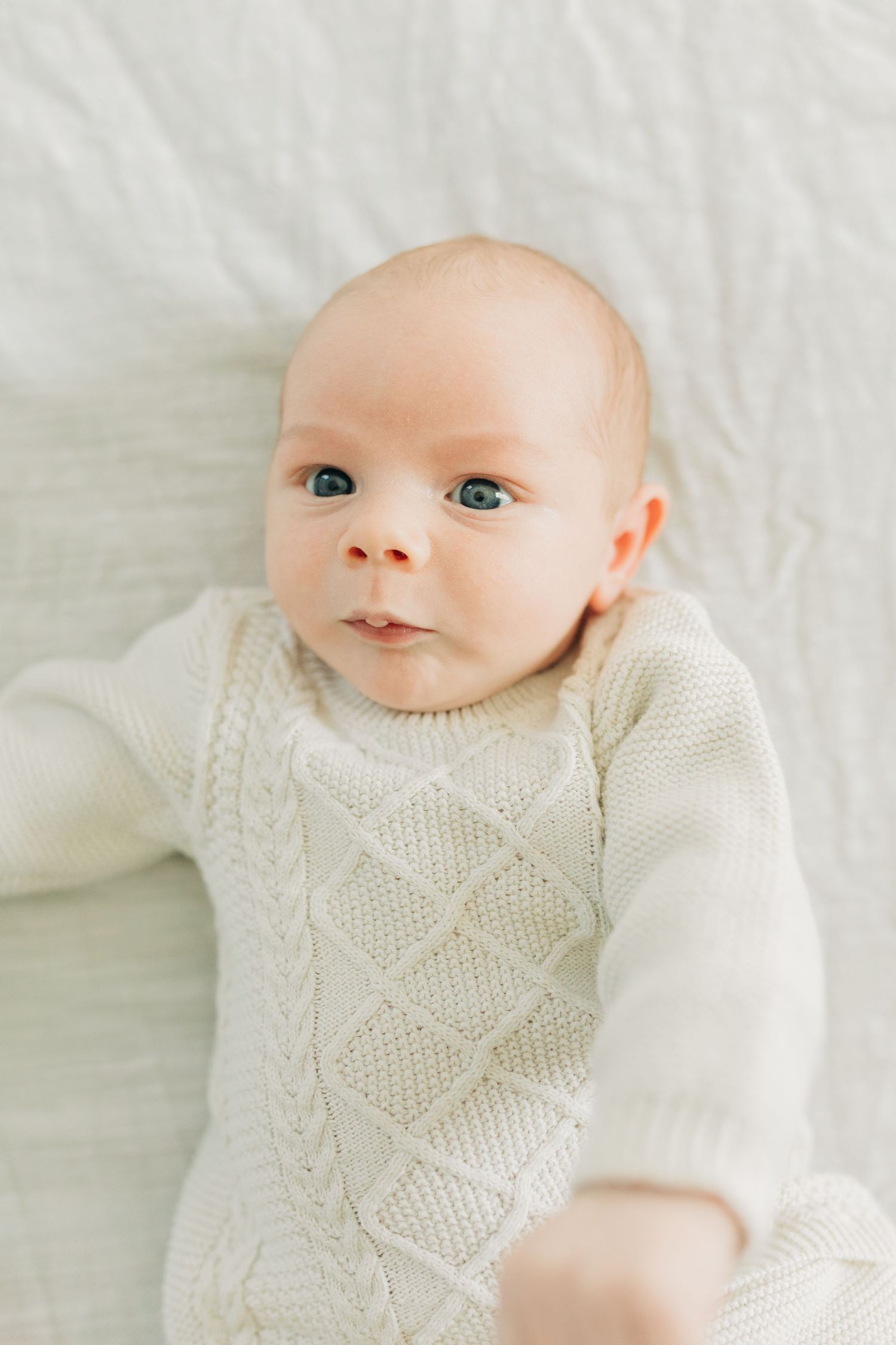 Close-up portrait of infant wearing cream knit sweater on white bedding.