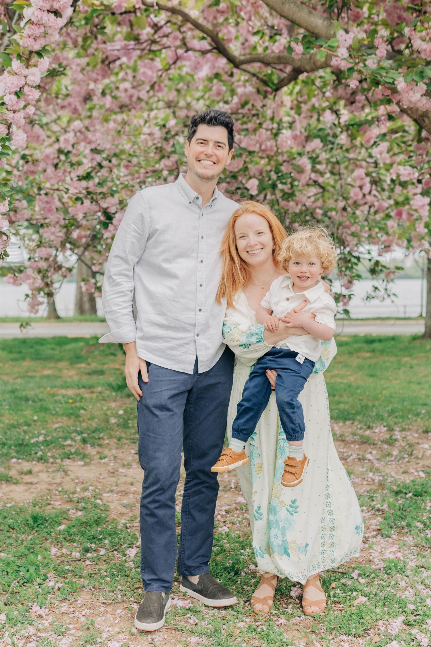 A family poses together under blooming pink cherry blossom trees in a spring garden setting.