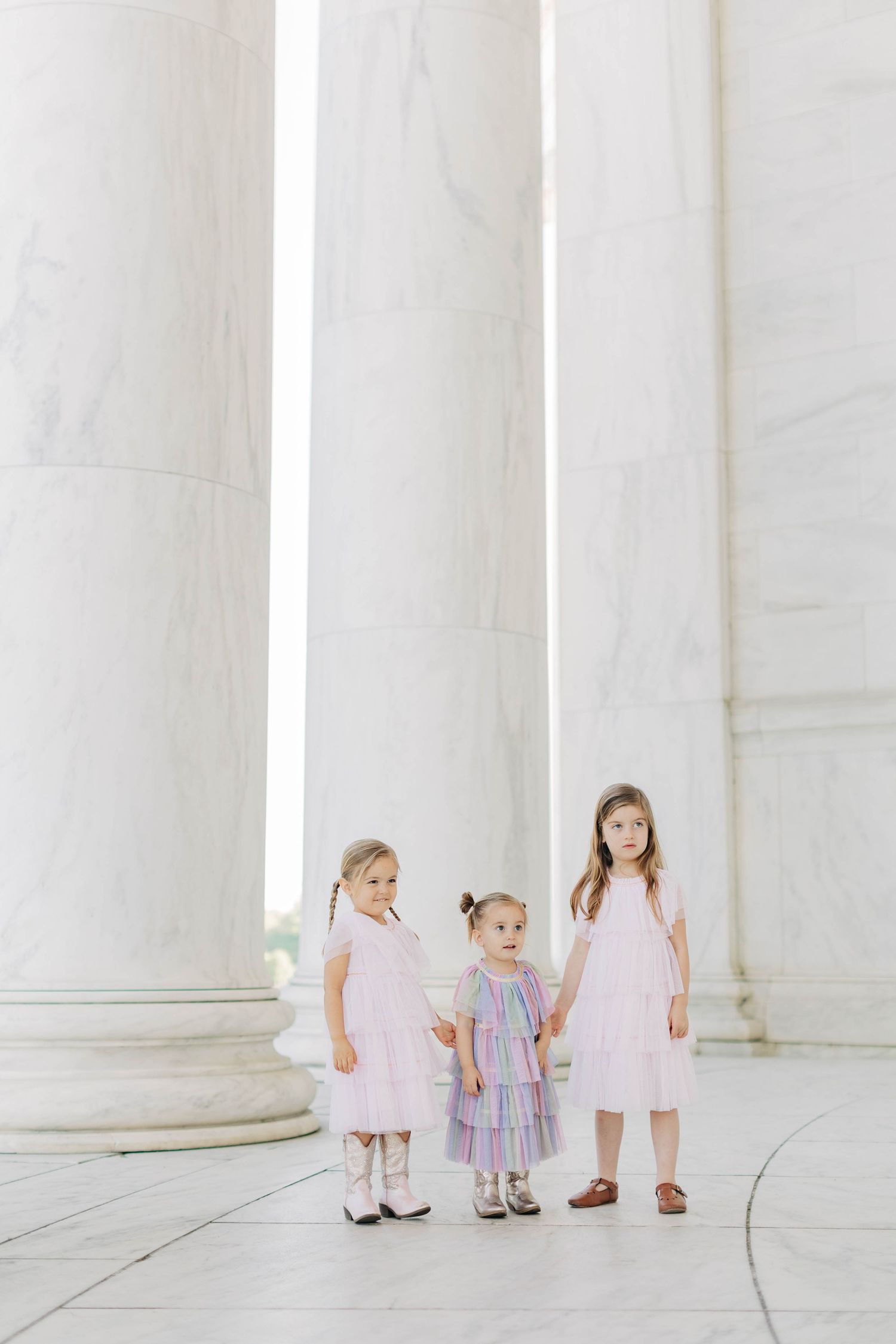 Three little girls in pastel dresses stand holding hands between tall white marble columns.