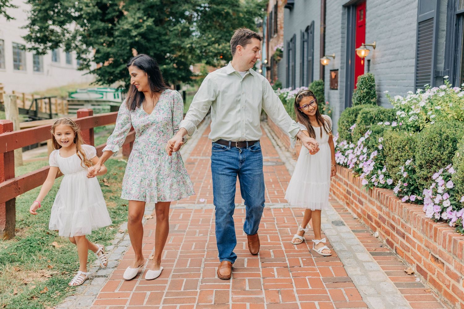 A happy family of four walks hand-in-hand on a brick path, with two girls in white dresses and parents in casual attire.