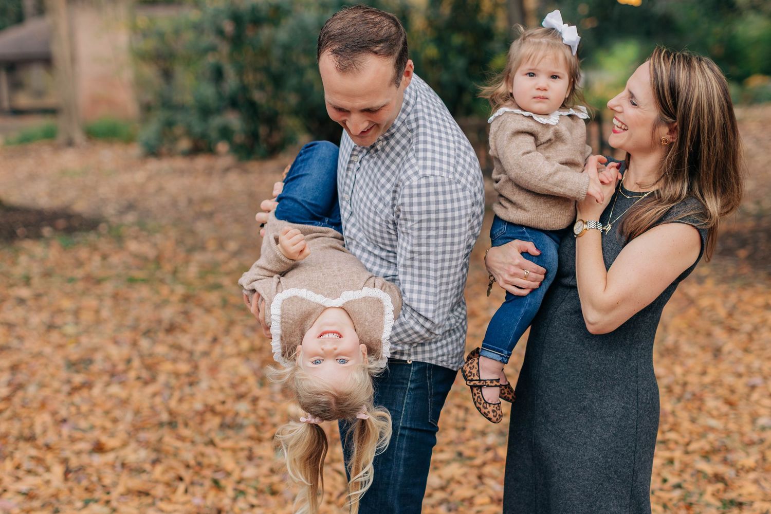 Family of four enjoying autumn outdoors, dad playfully flipping daughter upside down while mom holds toddler.