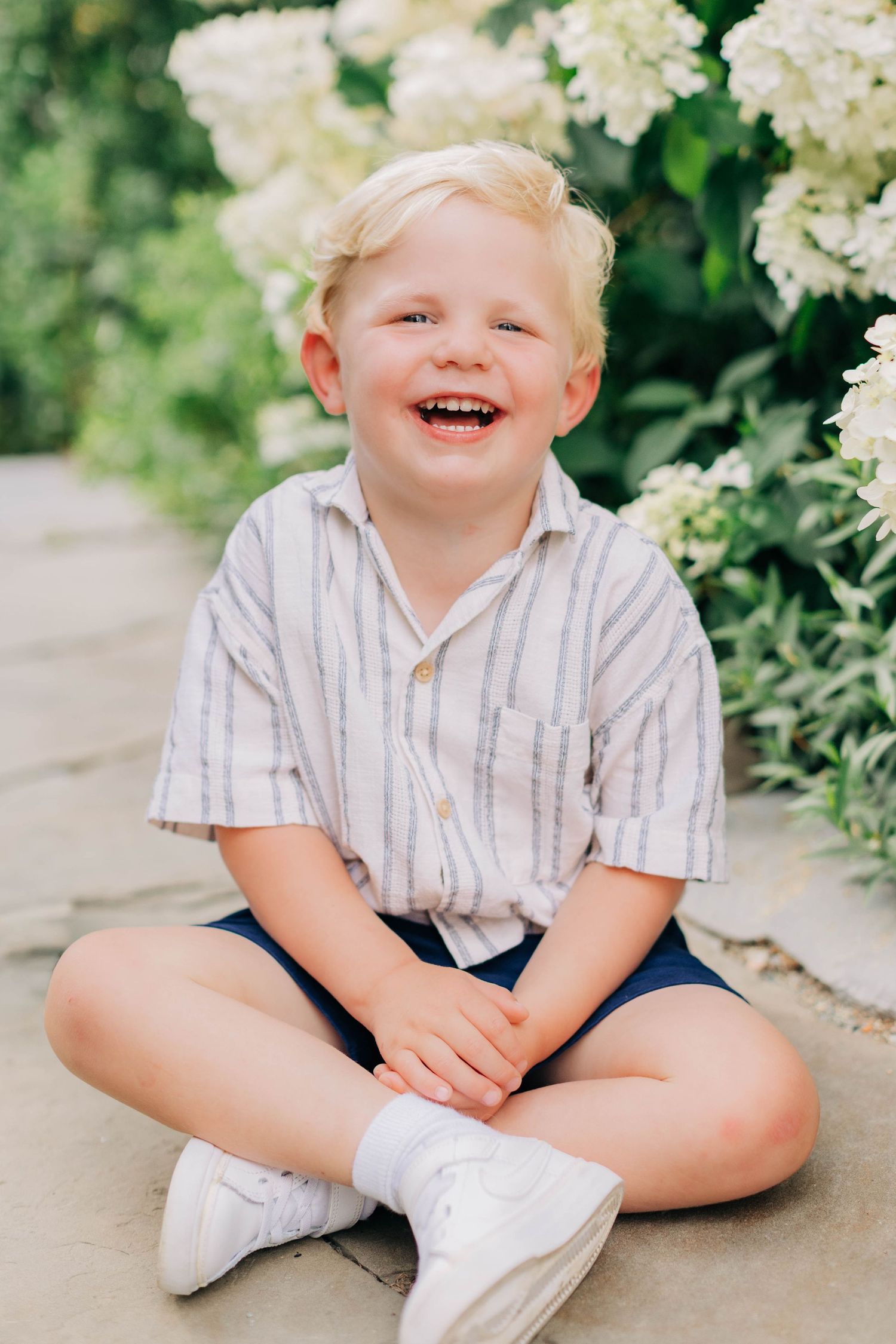 A cheerful blonde toddler boy sits cross-legged on a path, laughing joyfully, wearing a striped shirt and blue shorts.