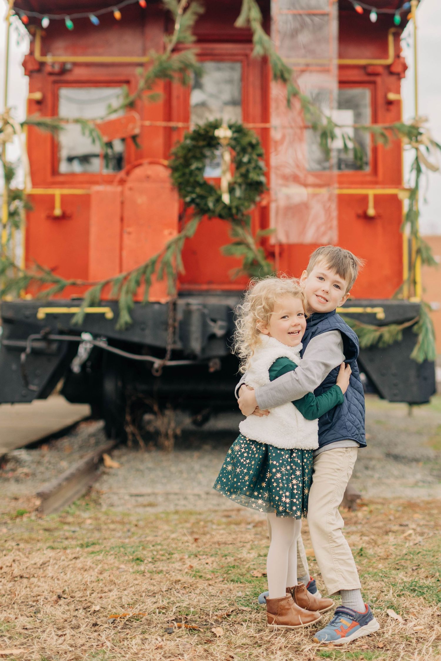 Two children hugging in front of a red Christmas-decorated train car with a wreath and holiday lights.