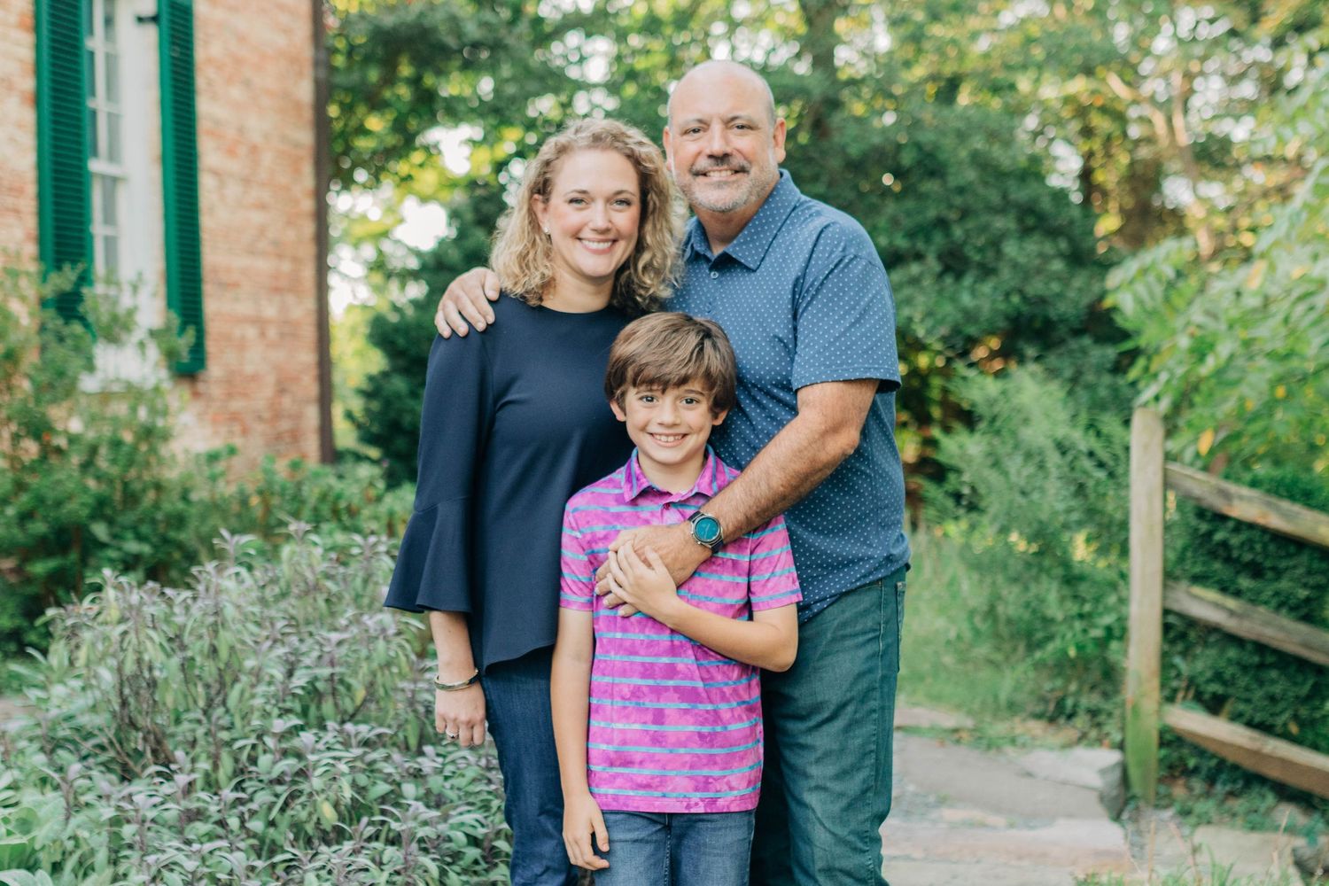 Family portrait taken outdoors with green trees and brick building in background.
