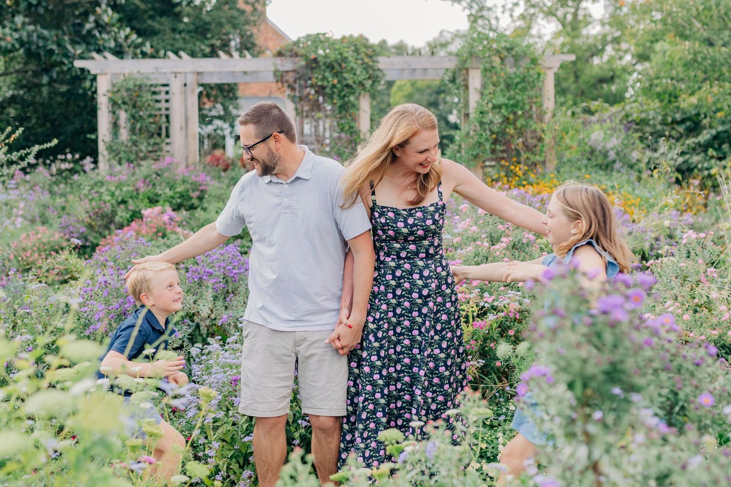 A family walks together through a blooming garden with purple flowers and a white pergola in the background.