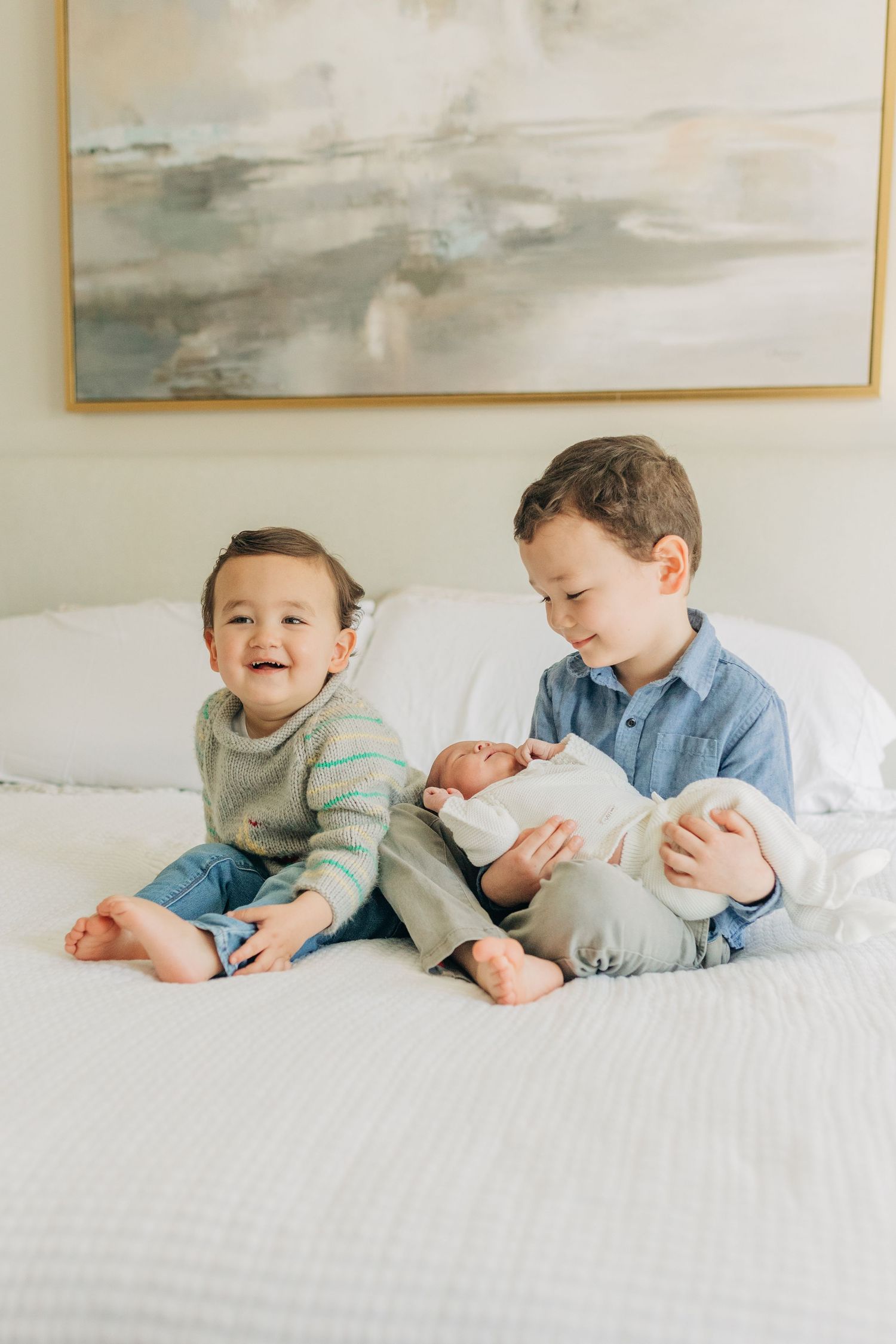 Three young children interact playfully on white bedding in a bright room.