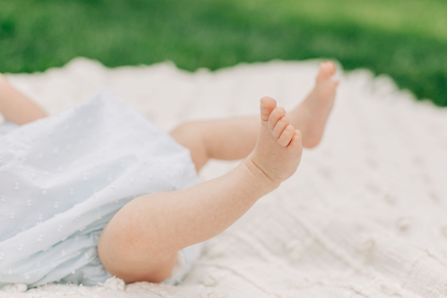 Tiny baby feet and legs on white blanket outdoors with grass in background.
