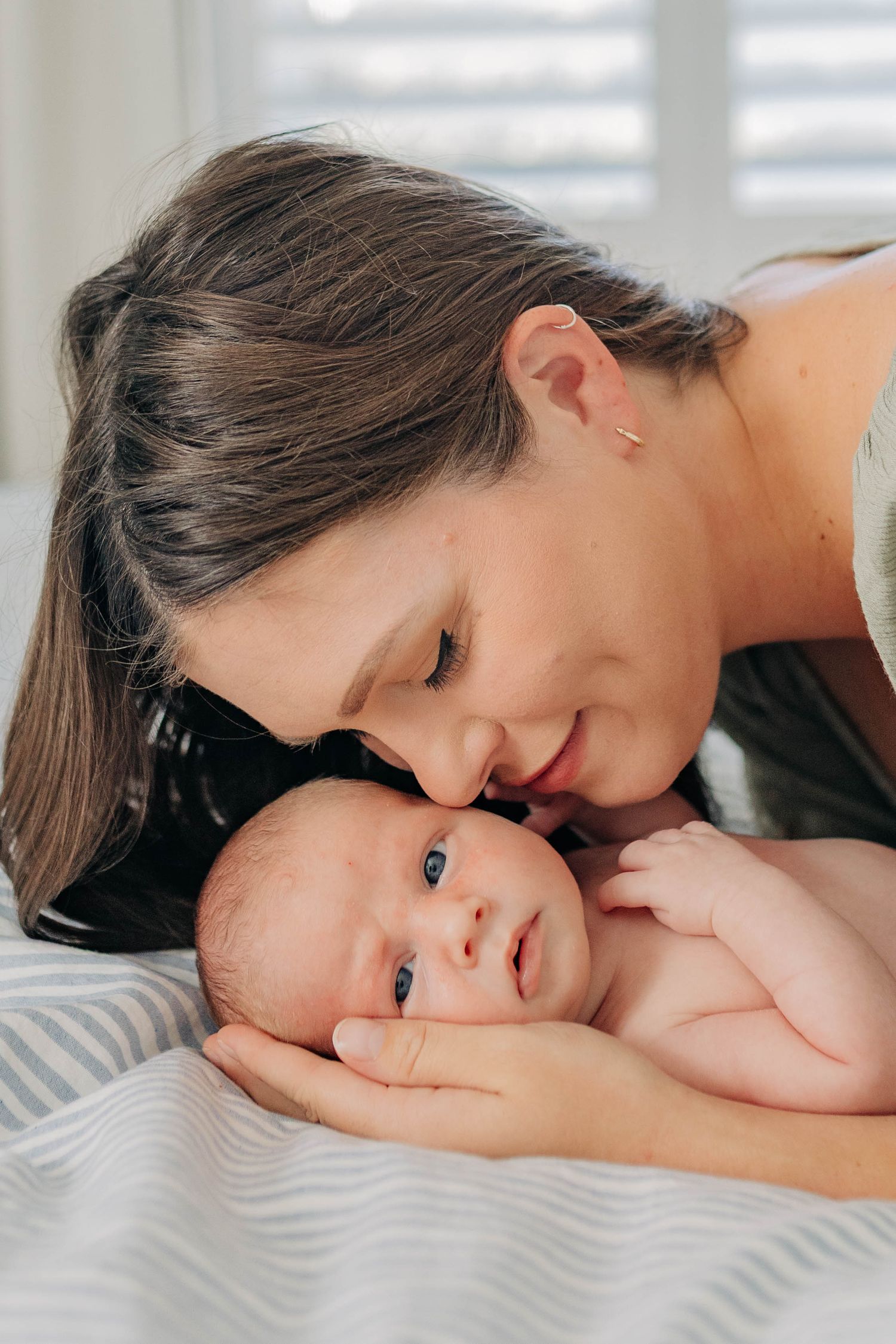 A tender moment between parent and baby lying on white bedding in natural window light.