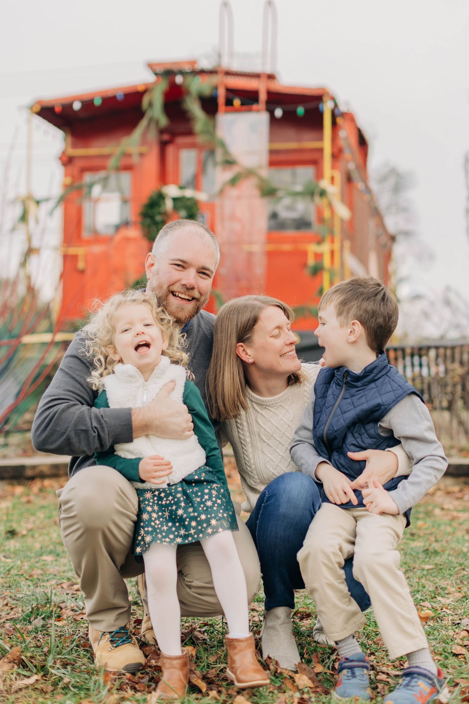 Happy family of four laughing together outdoors near a decorated red train car during the holiday season.