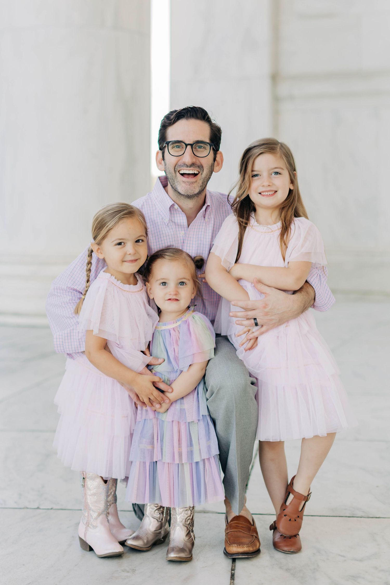 A smiling man with glasses poses with three young girls in pastel dresses at a marble monument for a family portrait.