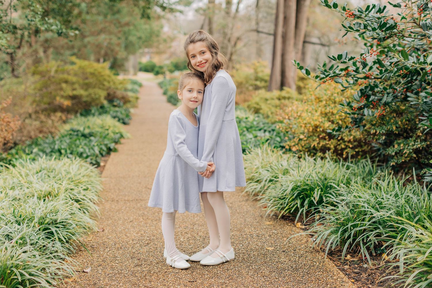 Two girls in matching gray dresses pose on a garden path surrounded by lush greenery.