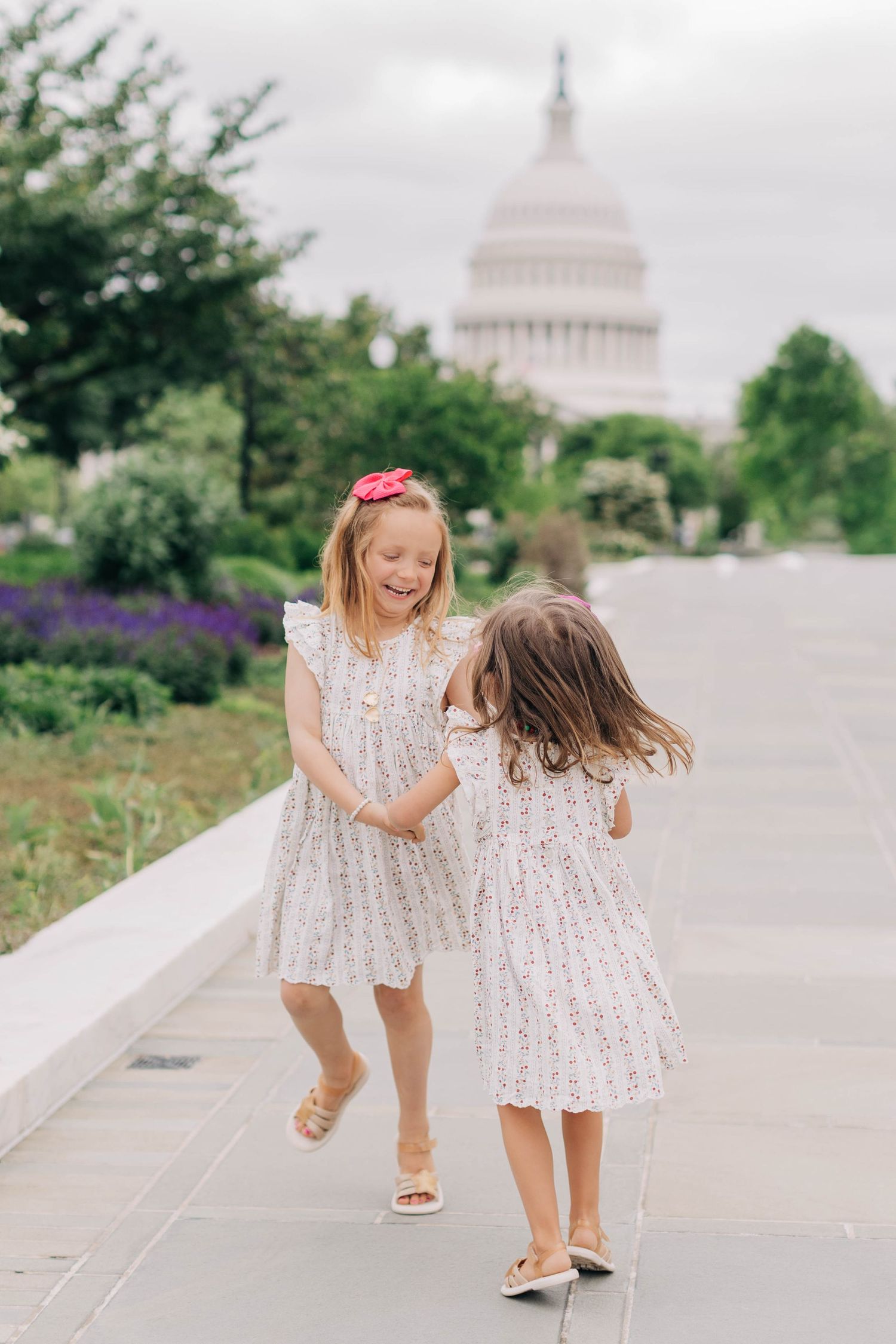 Two young girls in matching white floral dresses laugh and play on a path near the US Capitol building in Washington DC.