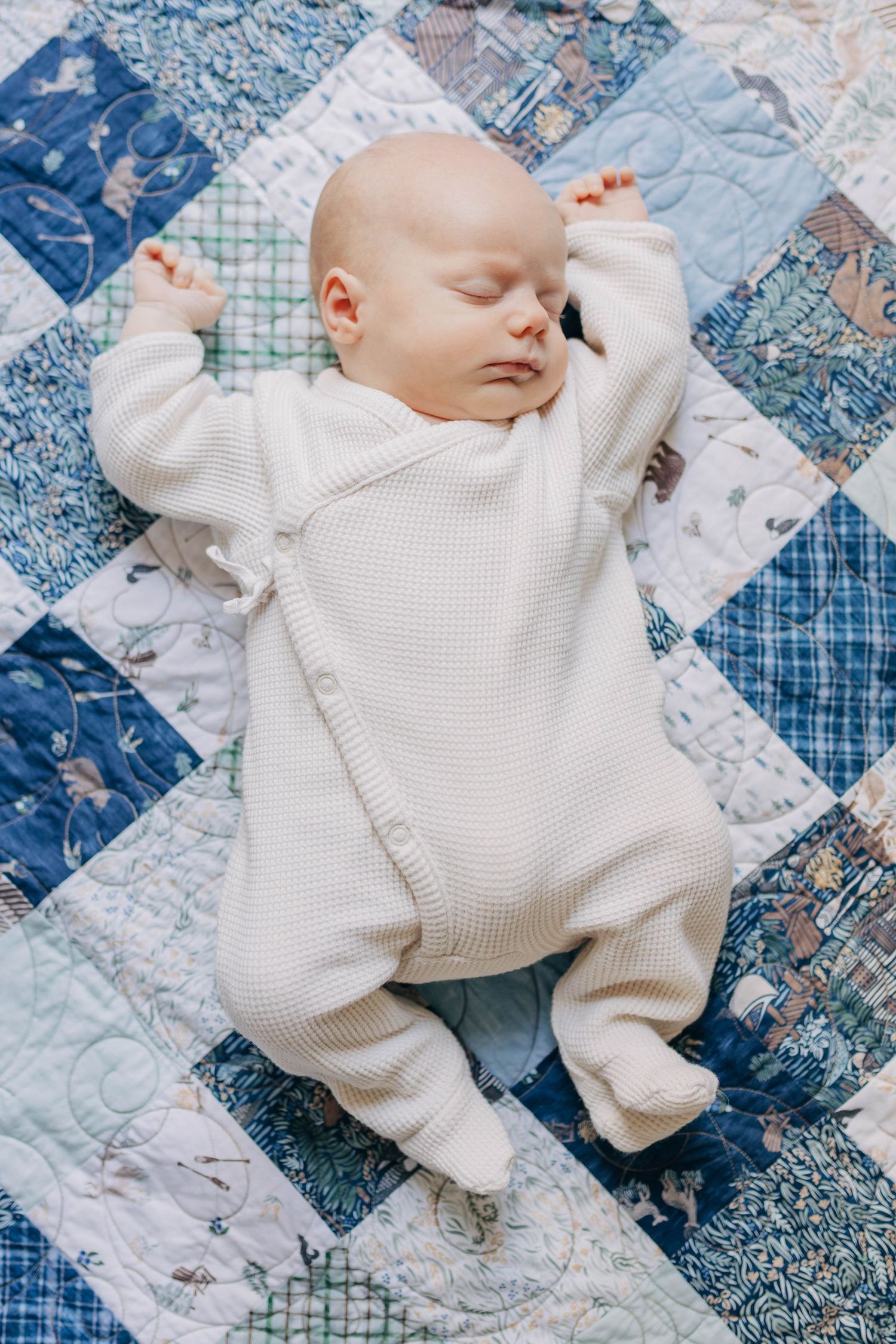 A peaceful newborn baby in a white knit onesie sleeping on a colorful patchwork quilt.