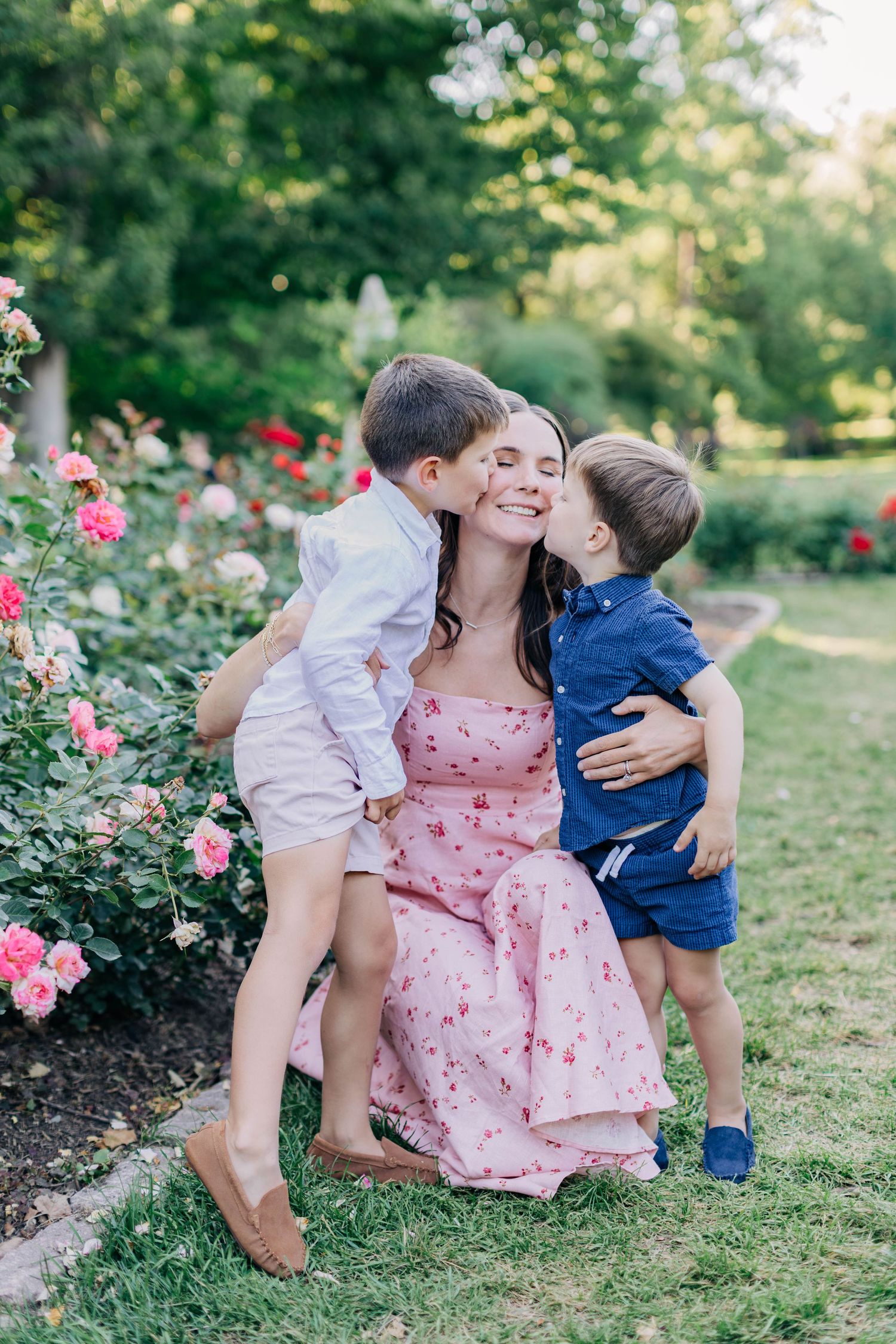A mother in a pink floral dress smiles as her two young sons kiss her cheeks in a beautiful rose garden.