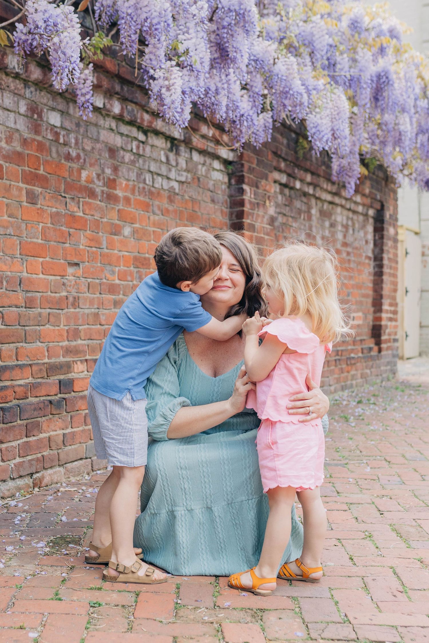 A mother kneels as her two young children kiss her cheeks near a brick wall with blooming wisteria.