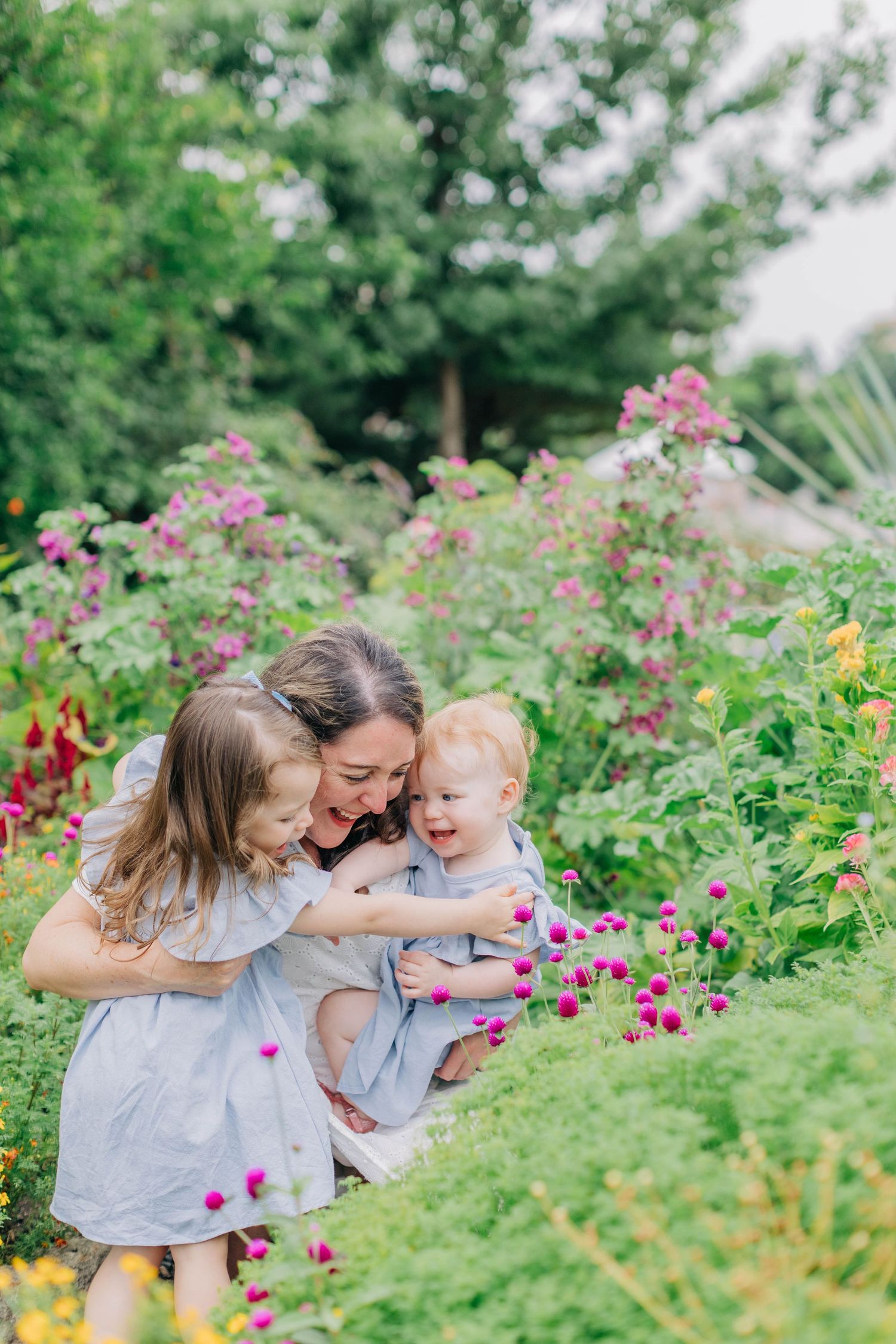 A mother lovingly embraces her two young children among vibrant blooming flowers in a lush garden.