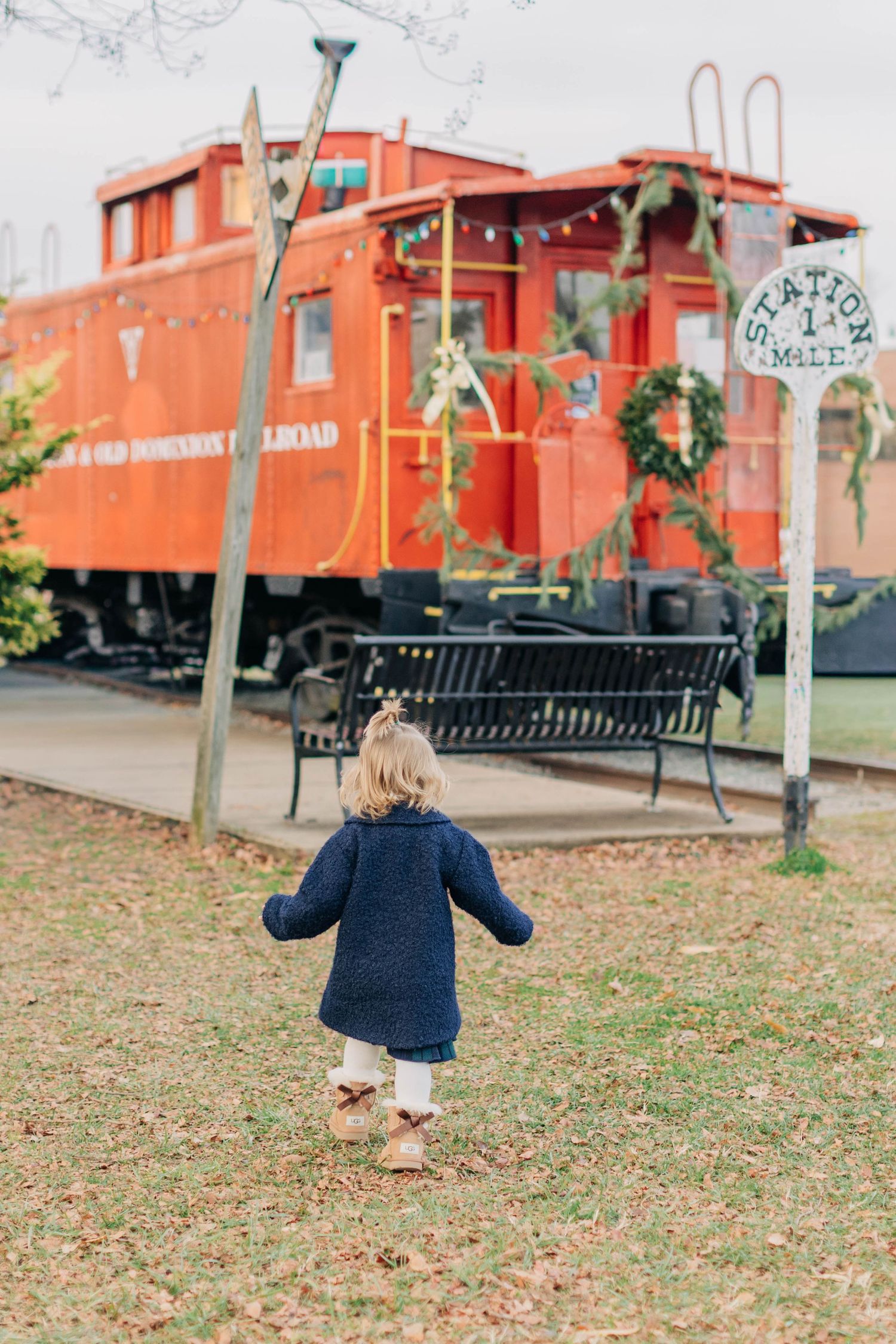 A toddler in a navy coat walks toward a red caboose train car decorated with a Christmas wreath at a railroad station.