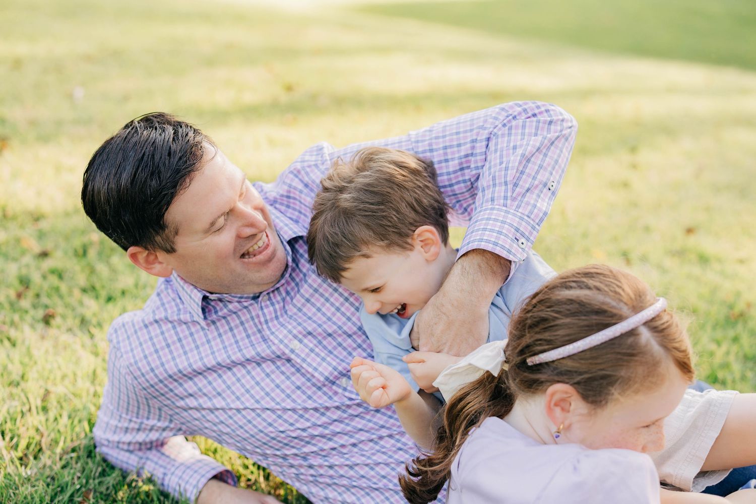 Father laughing with two young children while playing together on green grass outdoors.