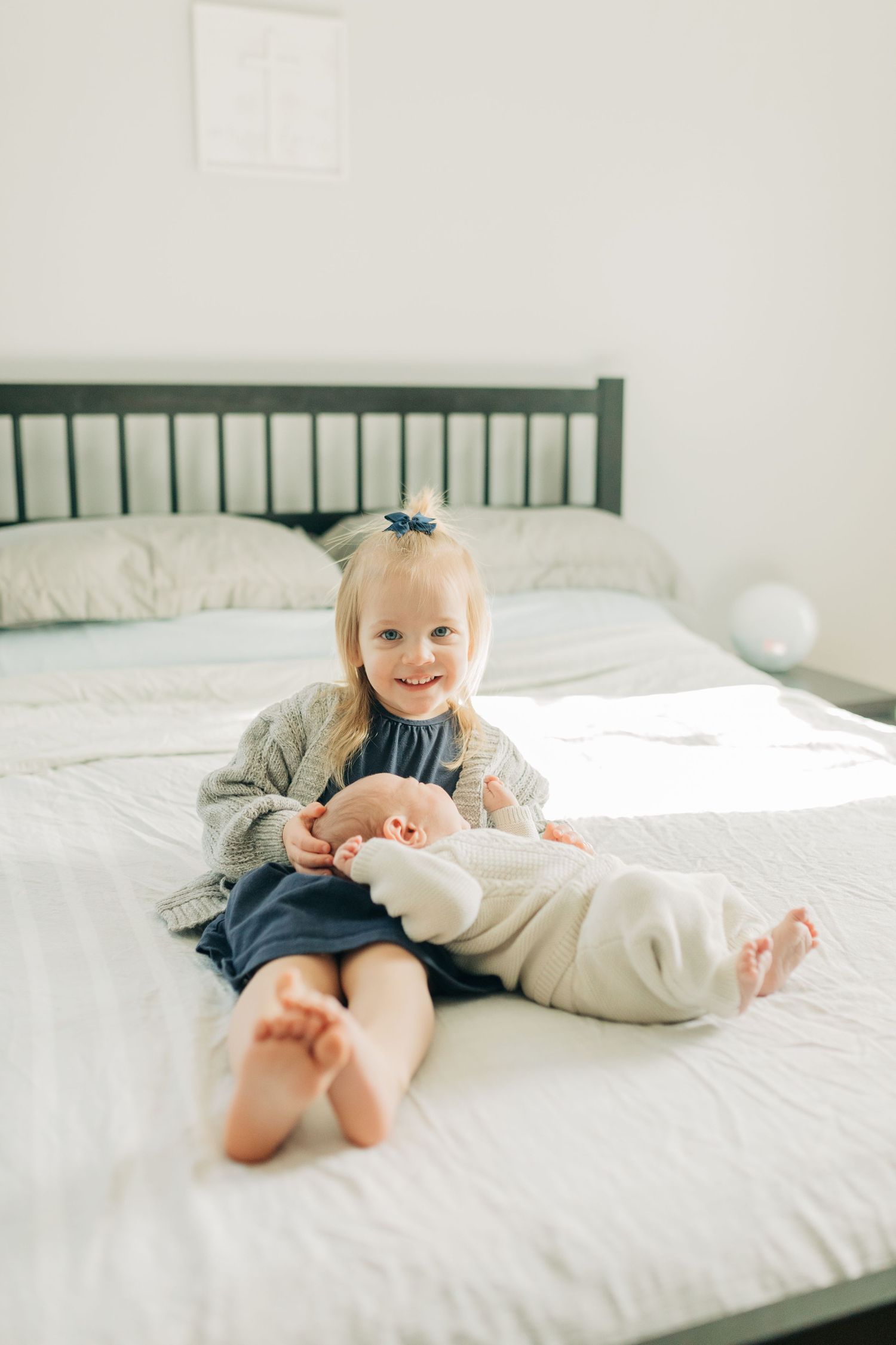 A young child sits with a baby on a white bed with green headboard.