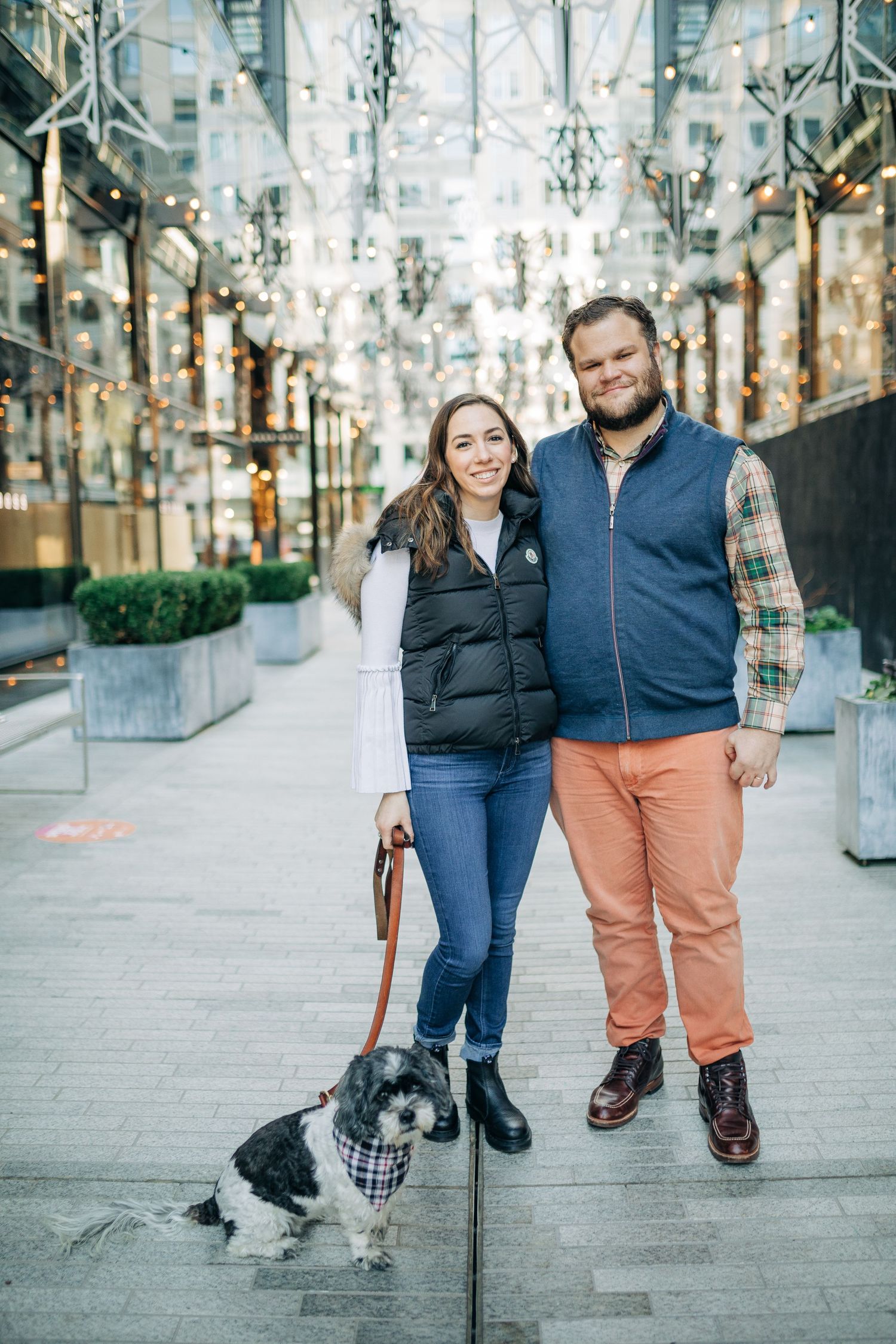 A couple walks with their small dog along a decorated urban pathway with twinkling lights.