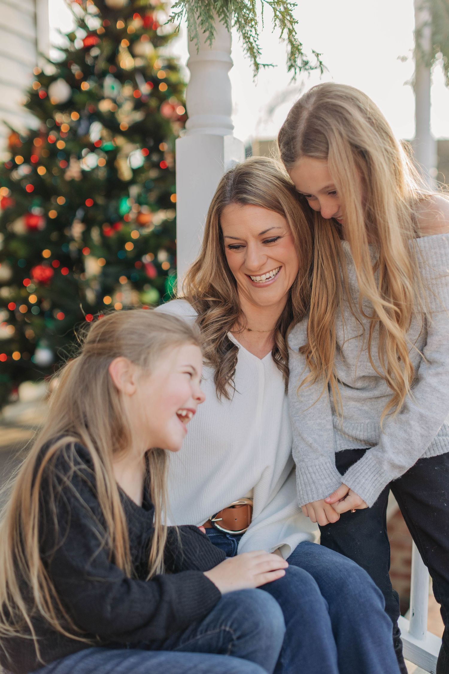 A mother and two daughters laugh together on a porch with a decorated Christmas tree in the background.