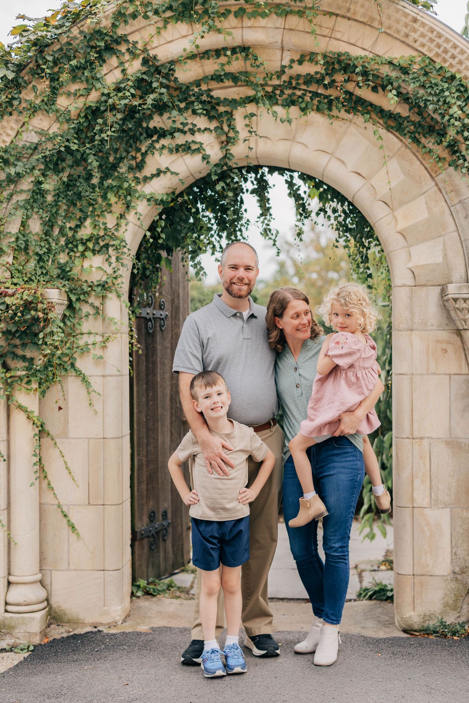 Smiling family of four poses together beneath an ivy-covered stone archway with a wooden gate, parents holding their young son and daughter.