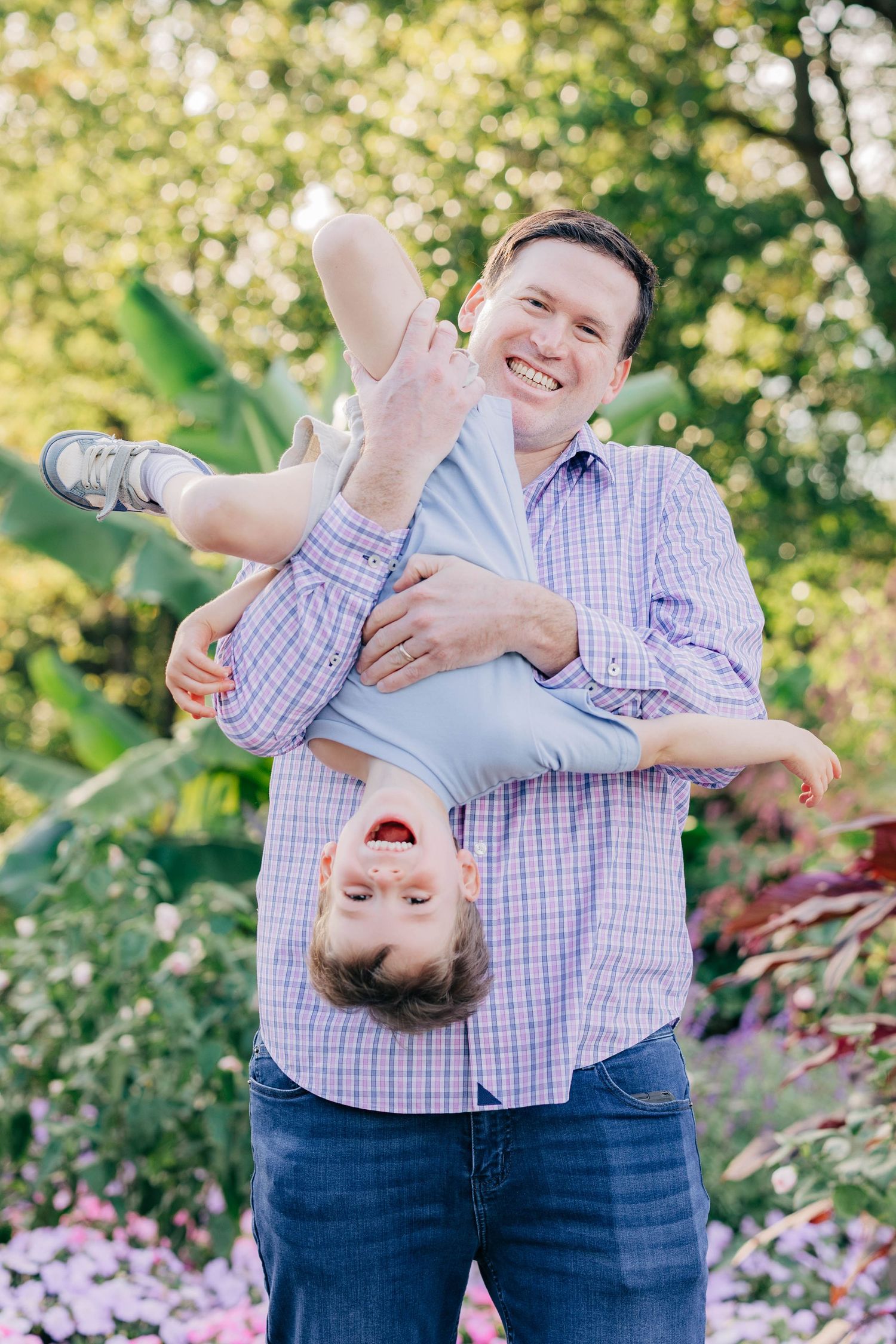 A smiling father holds his laughing child upside down in a lush garden, both wearing blue outfits.