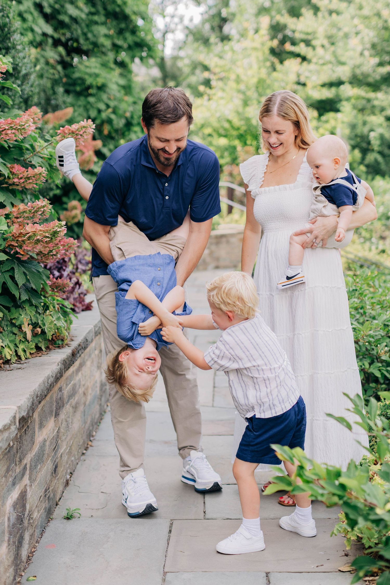 Happy family enjoying outdoor time, father playfully holding child upside down while mother holds baby in lush garden.