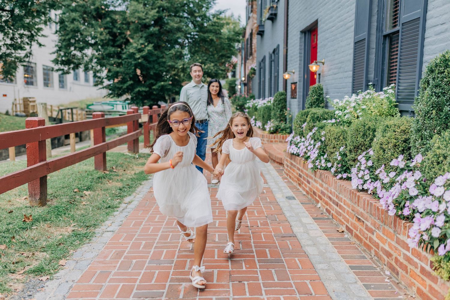 Two girls in white dresses run along a brick path while parents smile behind them near colorful flowers.