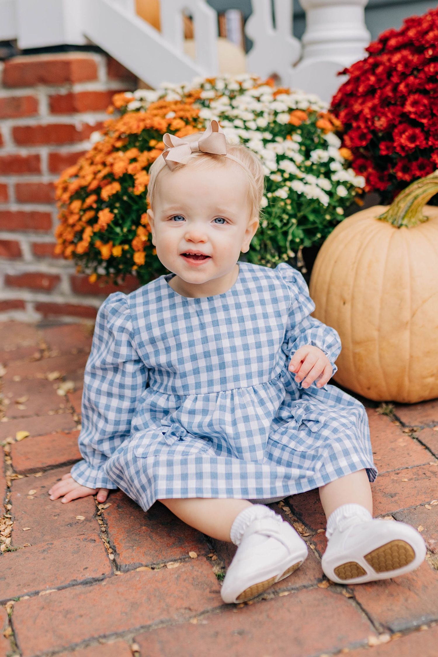 Smiling baby girl in blue gingham dress sitting on brick steps with fall mums and pumpkin decorations.