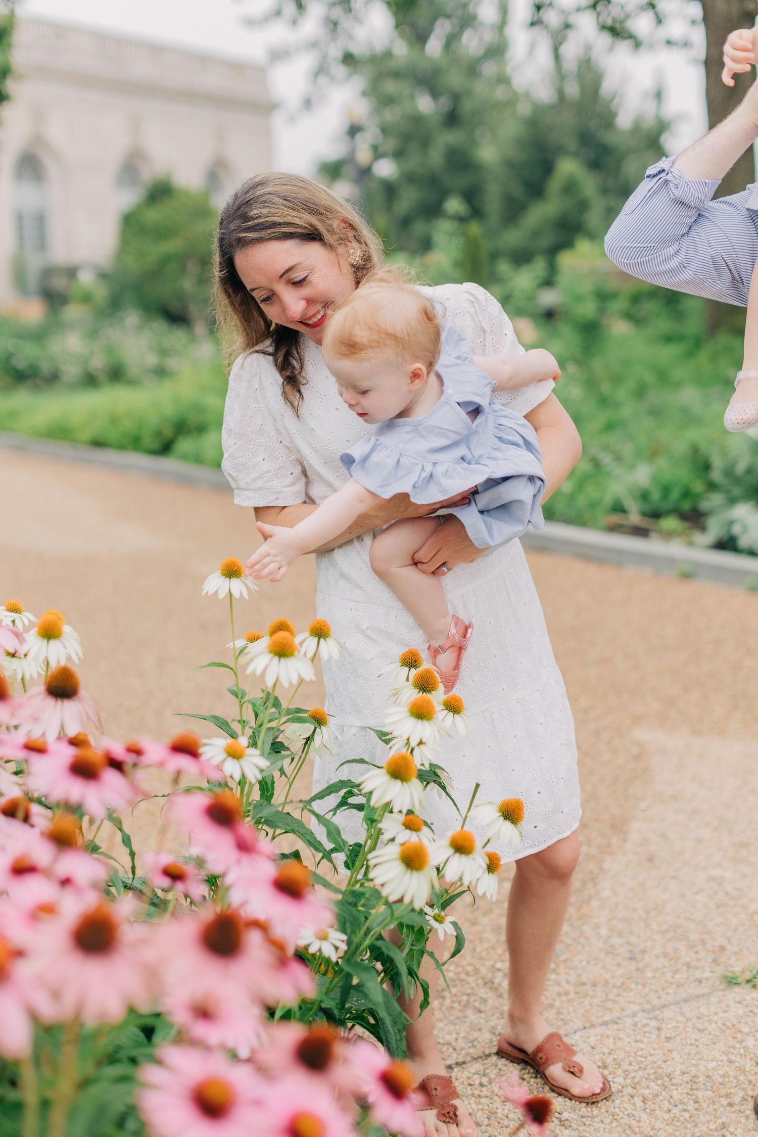 A smiling mother in a white dress holds her baby near blooming pink coneflowers in a lush garden.
