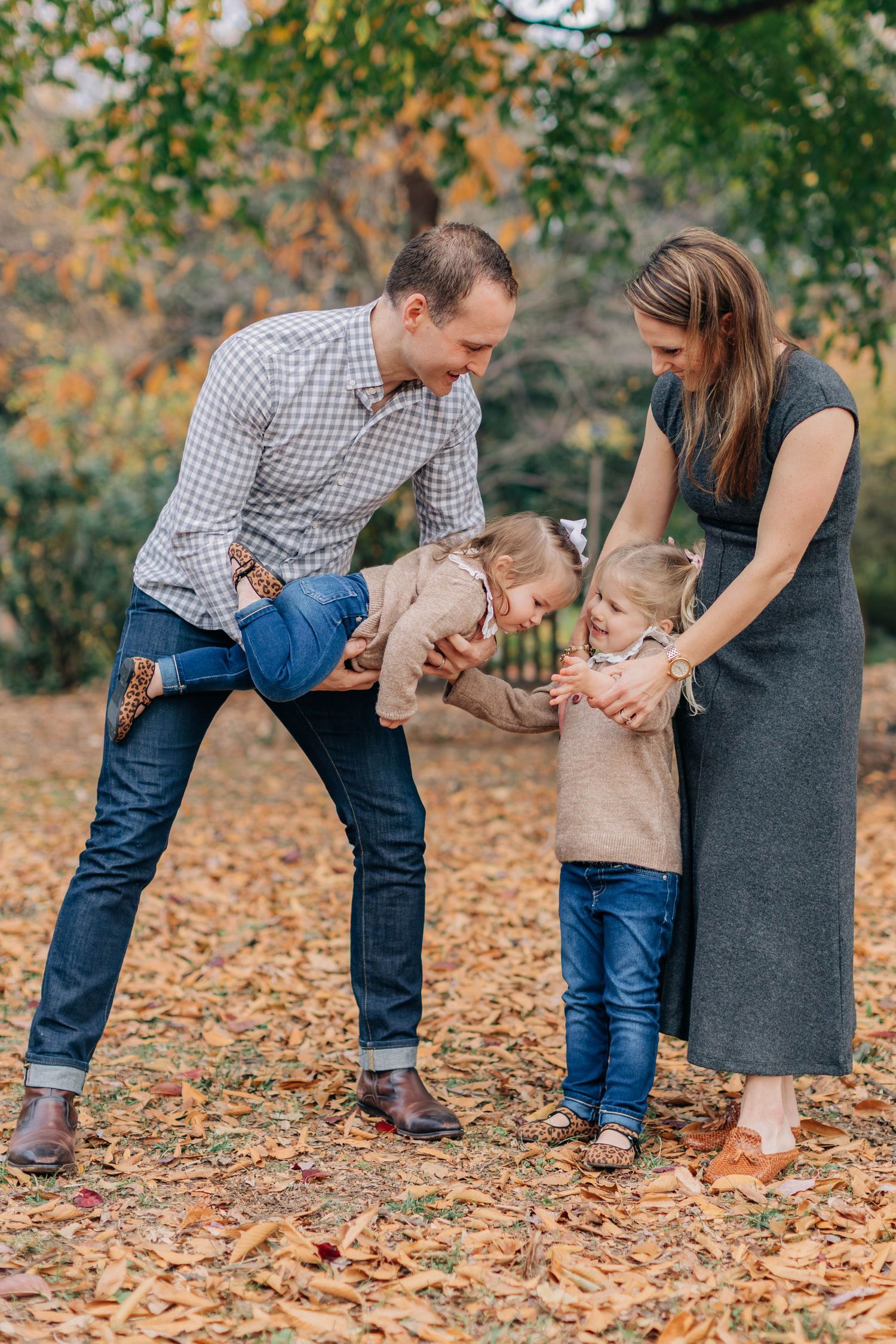 Family of four enjoying autumn park, father playfully lifting toddler while mother holds young daughter among fallen leaves.